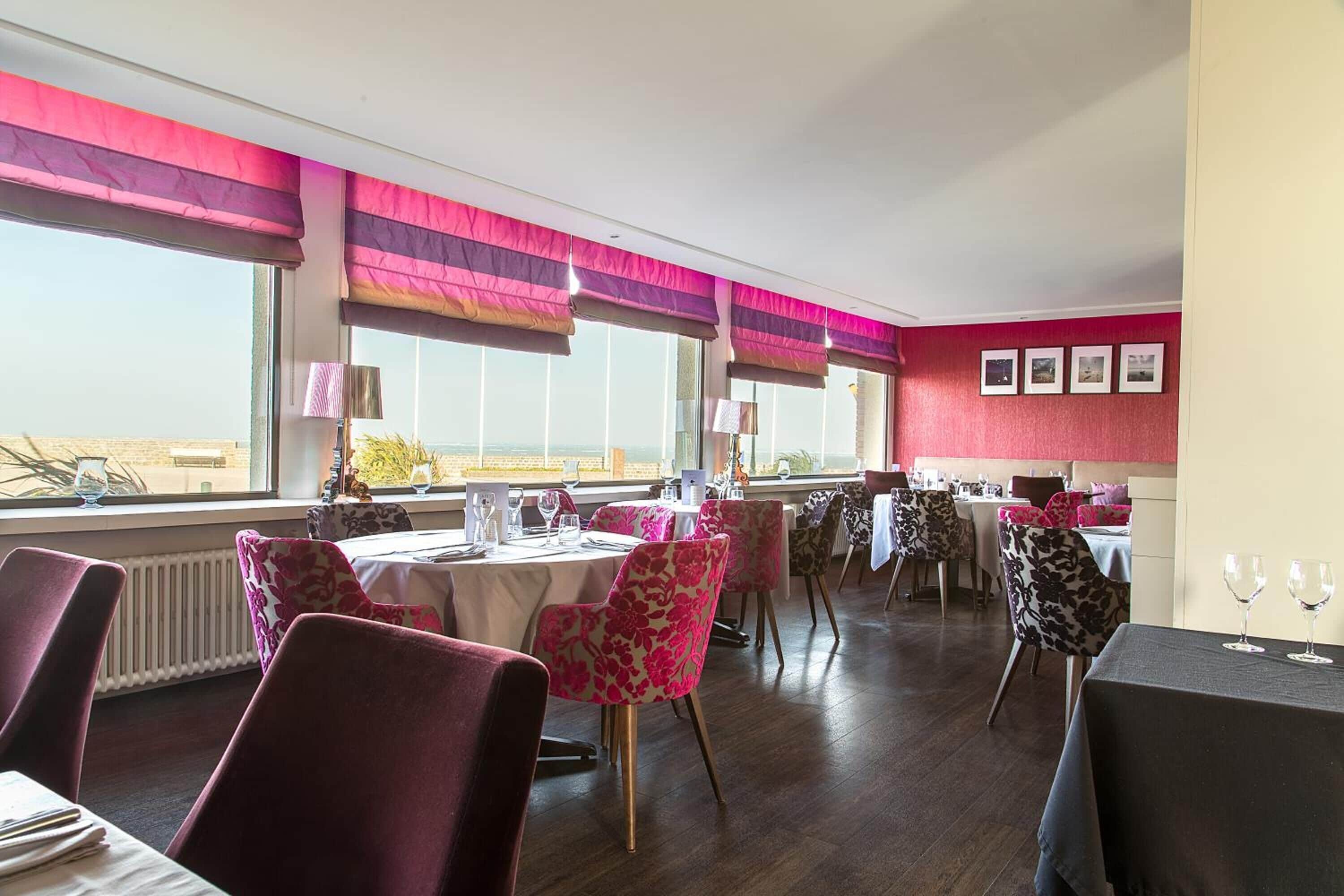 Un comedor con mesas dispuestas para el servicio, sillas con tapicería floral y amplias ventanas con cortinas a rayas.