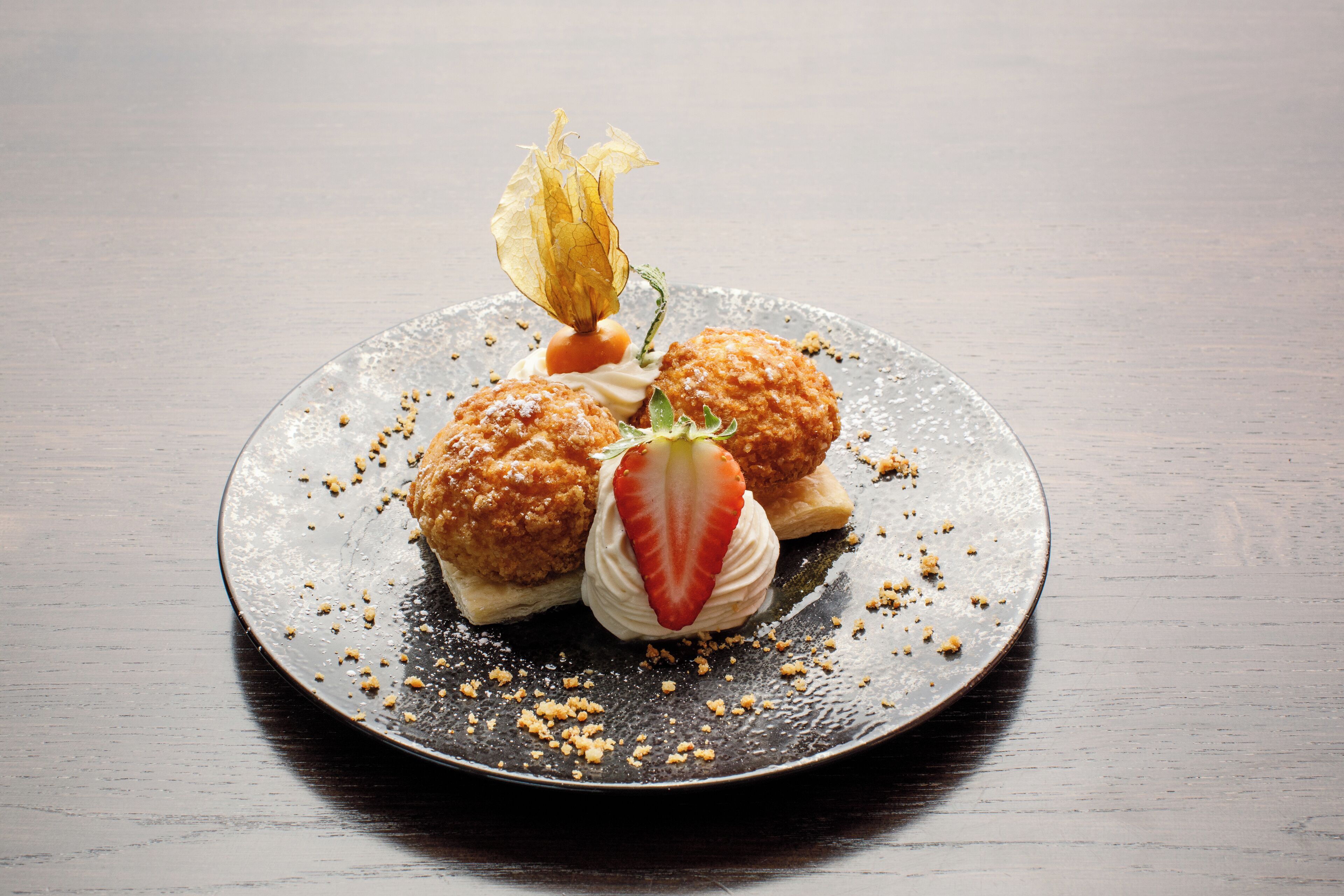 Un plato de postre con tres pastas redondas y doradas, una fresa y una flor decorativa sobre un plato oscuro.