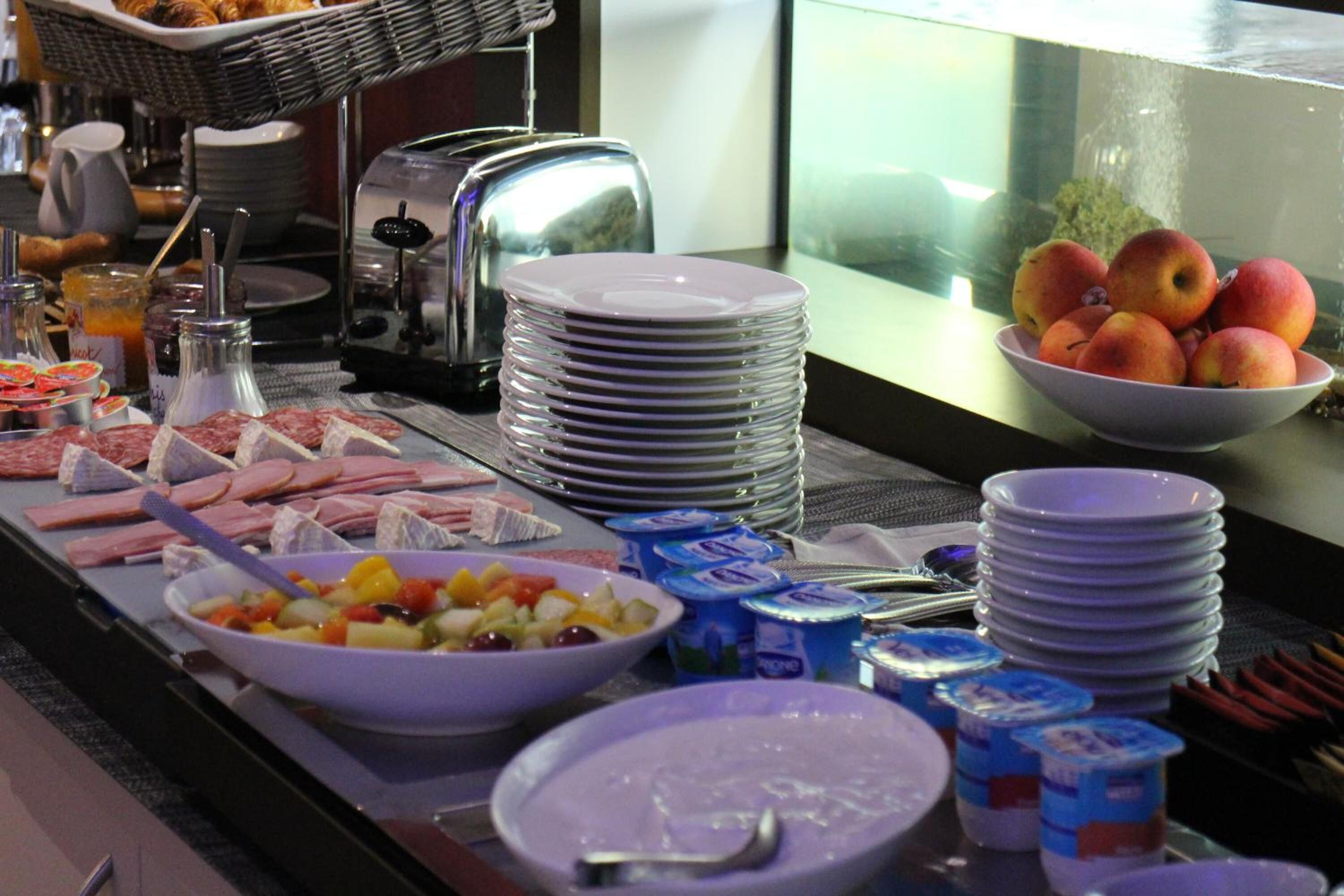 Montaje de bufé con diversos alimentos, como frutas, carnes y productos lácteos, dispuesto sobre una mesa con platos apilados y utensilios.