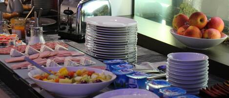 Montaje de bufé con diversos alimentos, como frutas, carnes y productos lácteos, dispuesto sobre una mesa con platos apilados y utensilios.