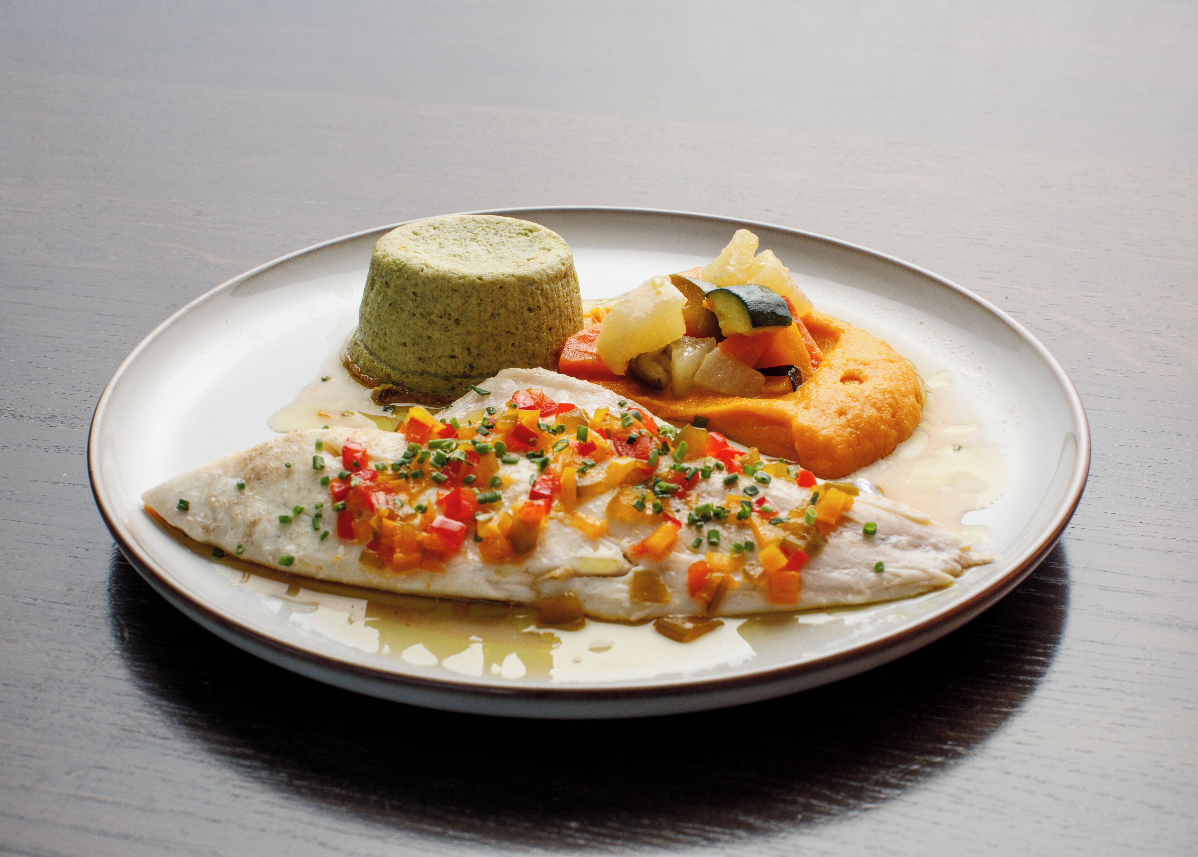 Un plato con un trozo de pescado, una porción de comida verde y un alimento horneado con verduras.