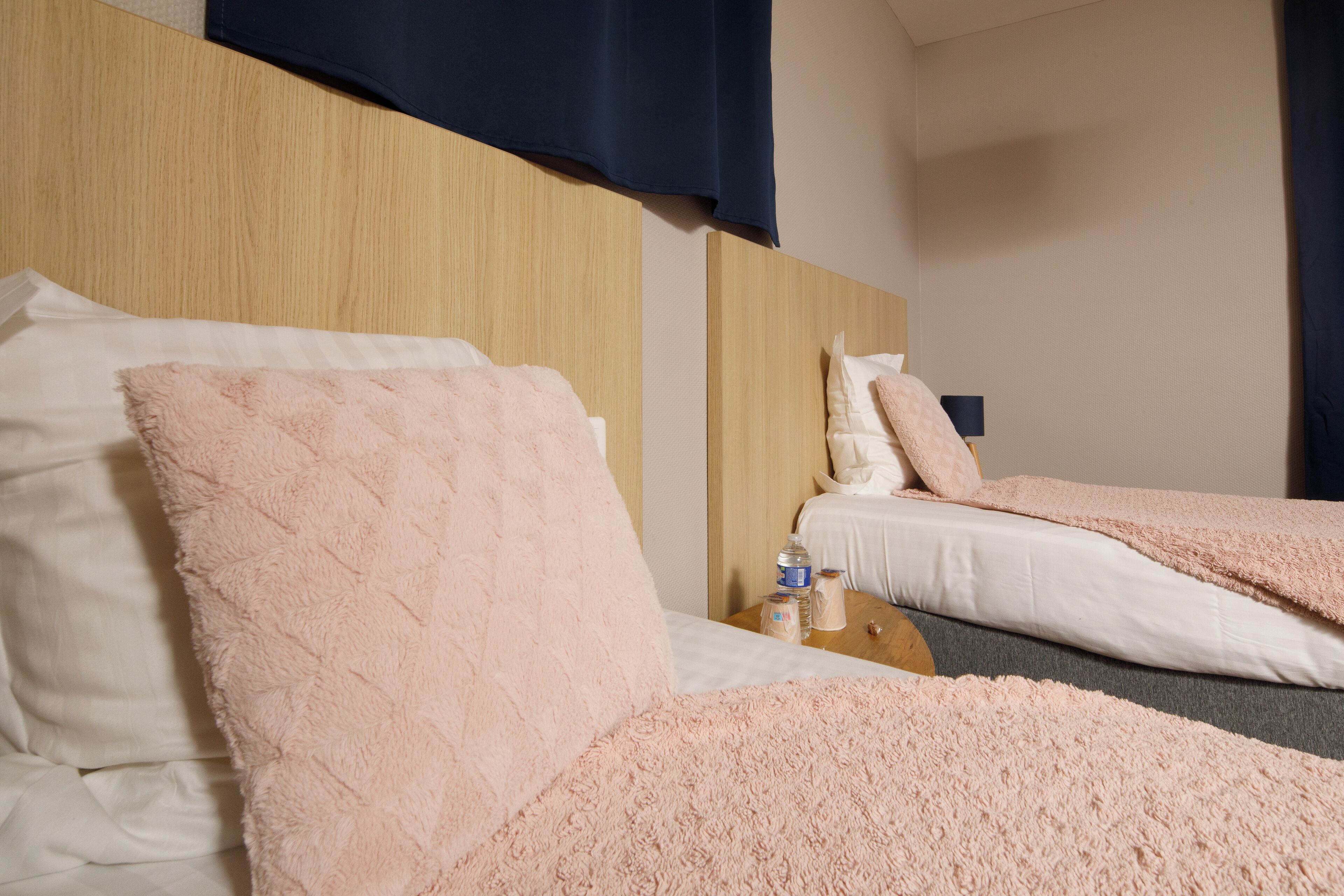 A hotel room with two beds, each with a fluffy pink blanket and white linens. There is a wooden headboard, a small table with a bottle and a tissue box, and a dark curtain on the window.