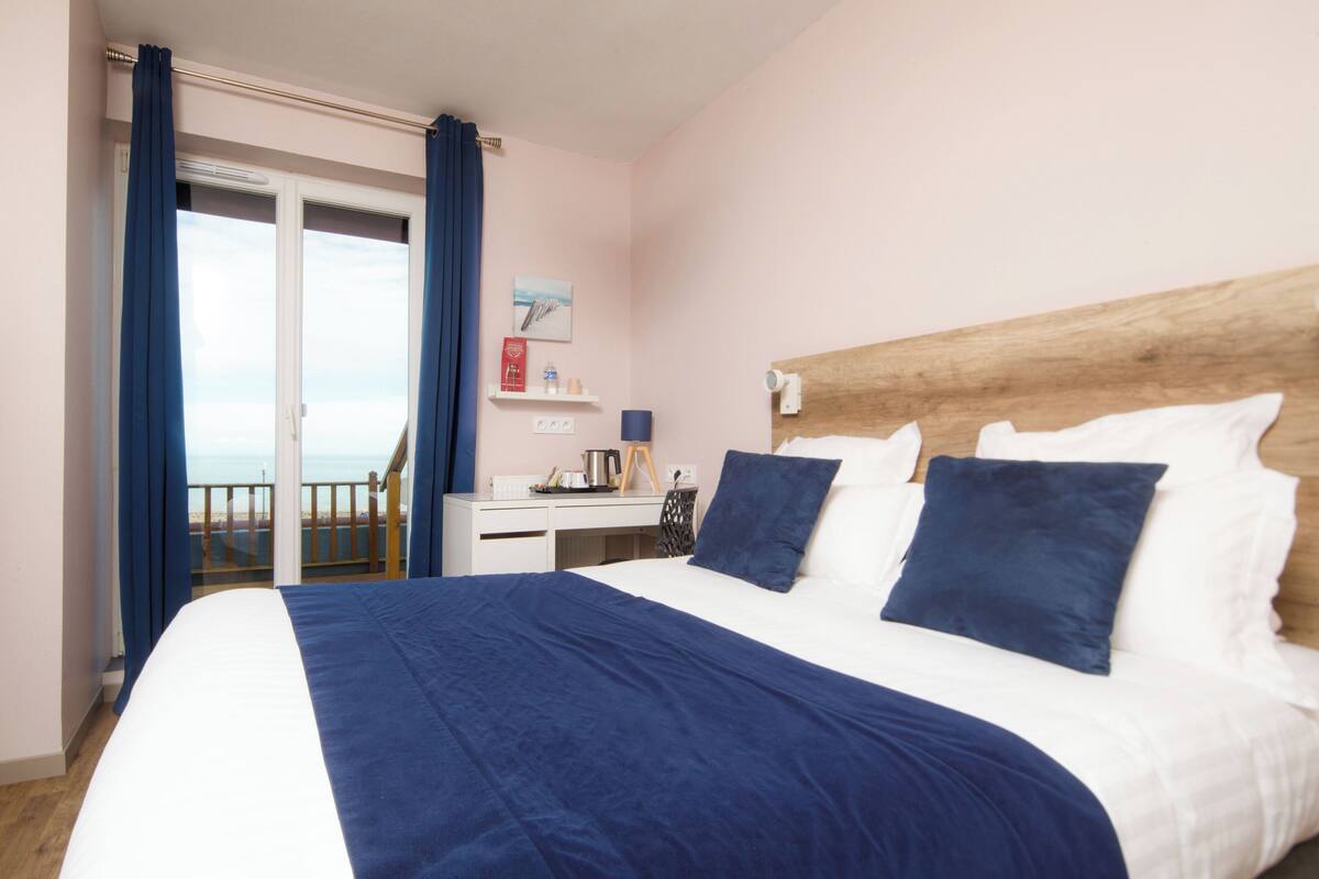 Hotelkamer met een groot bed, een houten hoofdeinde, een bureau met stoel, een klein tafeltje en uitzicht op het strand door een schuifdeur.