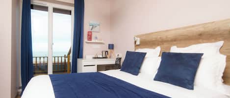 A hotel room with a large bed, a wooden headboard, a desk with a chair, a small table, and a view of the beach through a sliding door.