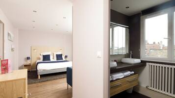 A modern hotel room with a bed, bedside table, and a view of the city through a large window.