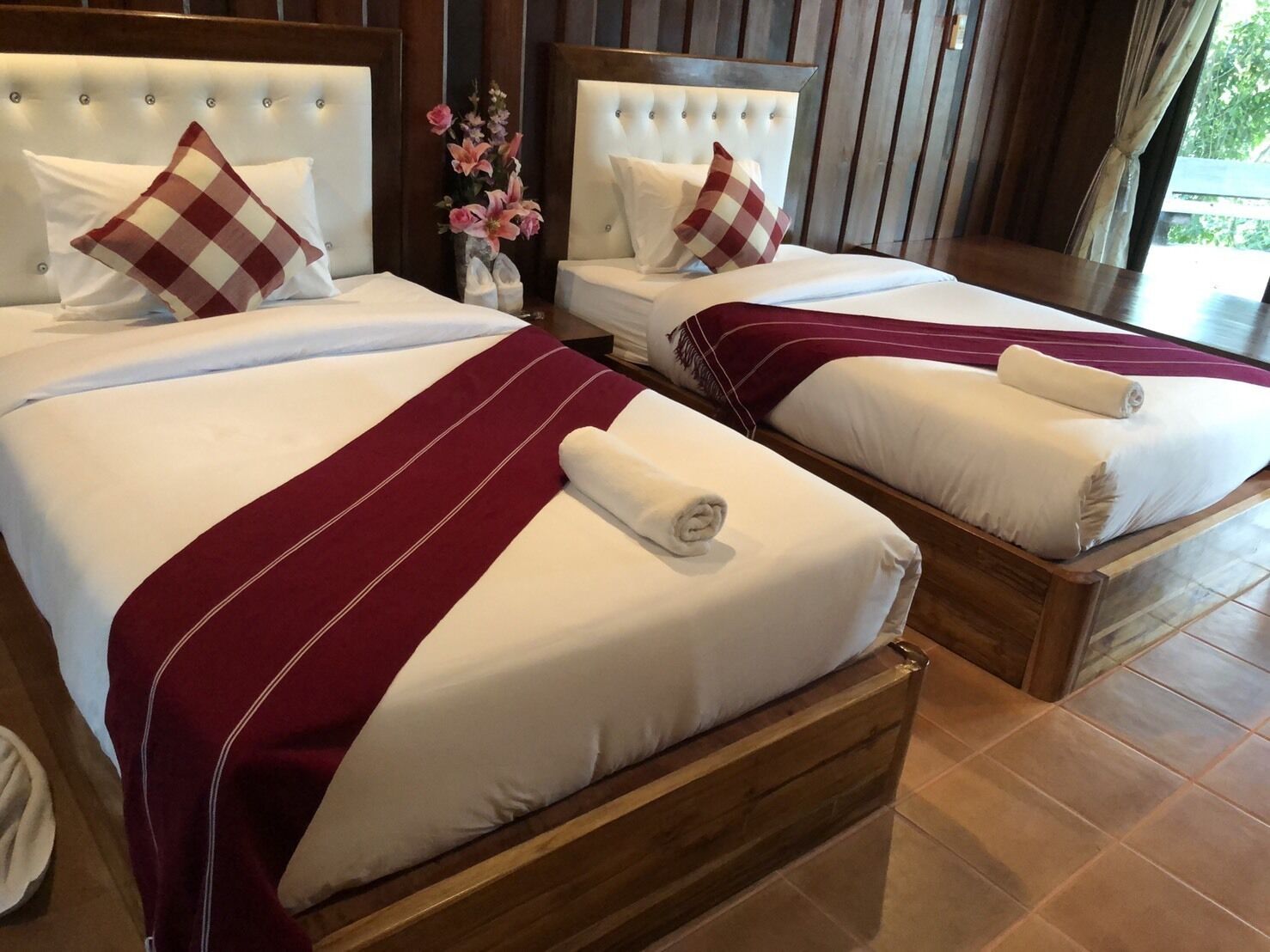 Two double beds with white linens and red and white checkered pillows, wooden headboards, and a vase with pink flowers.