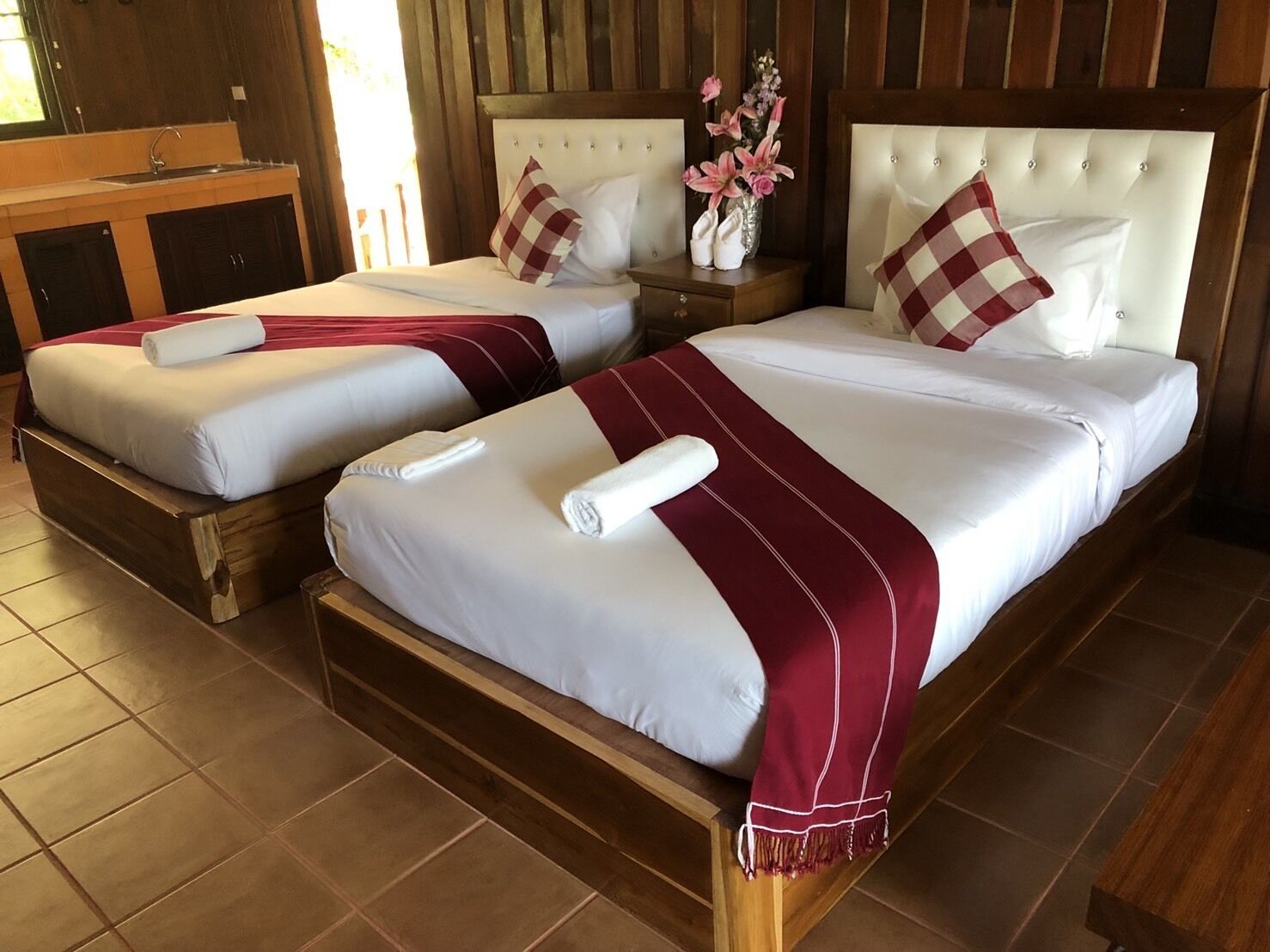 Dos camas dobles con ropa de cama blanca y detalles rojos, cabeceras de madera y una mesita de noche con un florero.