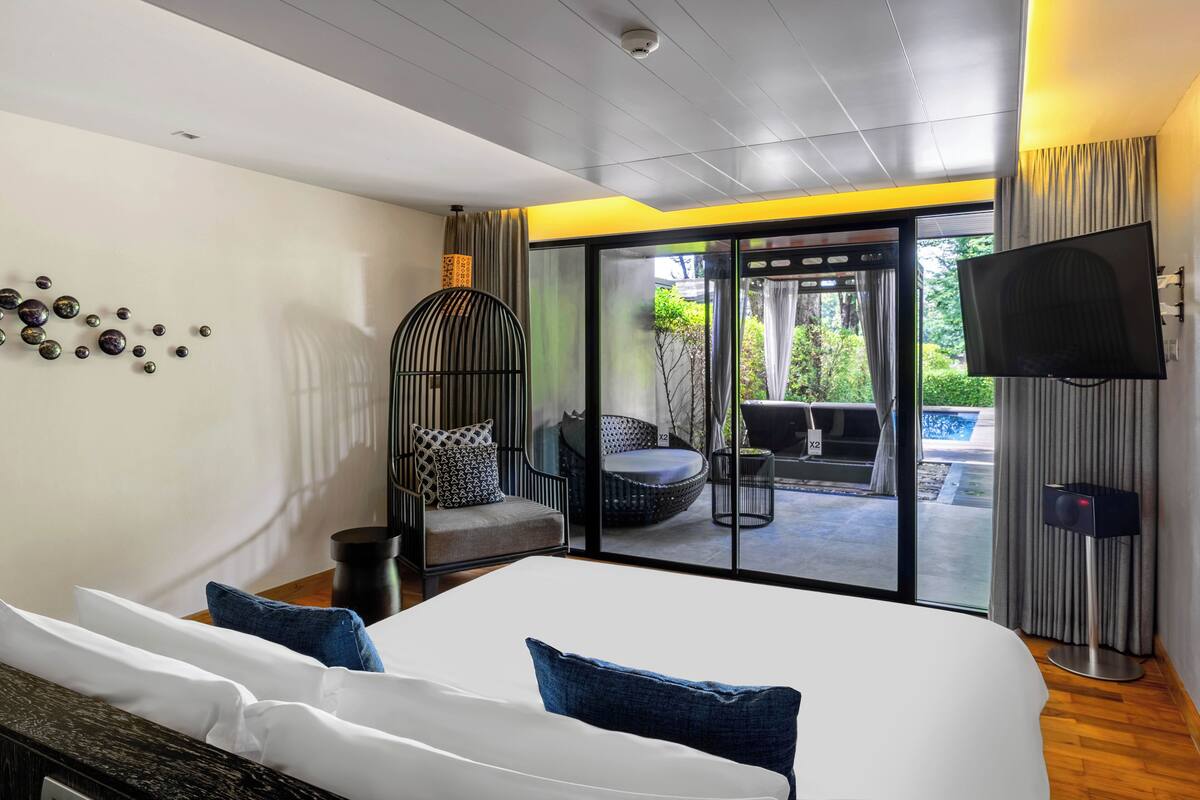 A modern hotel room with a large bed, a television, a balcony with outdoor seating, and a ceiling-mounted security camera.