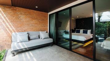A modern bedroom with a large sliding glass door leading to a balcony, a sofa in the adjacent living area, and a brick wall.