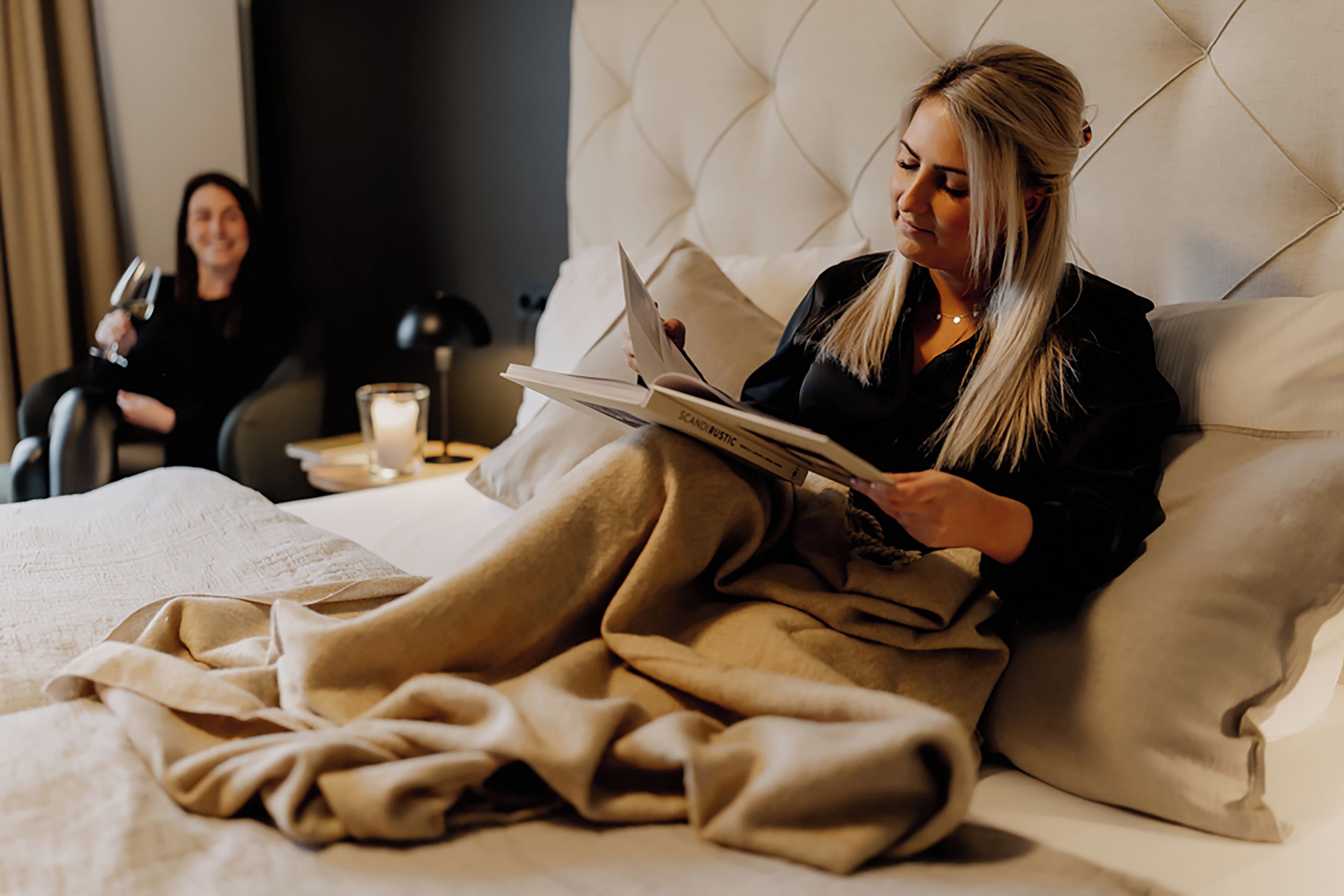 Una persona leyendo un libro en un dormitorio con una cama, almohadas y una mesita de noche con una vela.