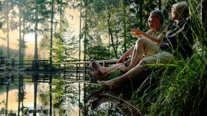 Two people sitting by a calm body of water, surrounded by trees and a wooden bridge.