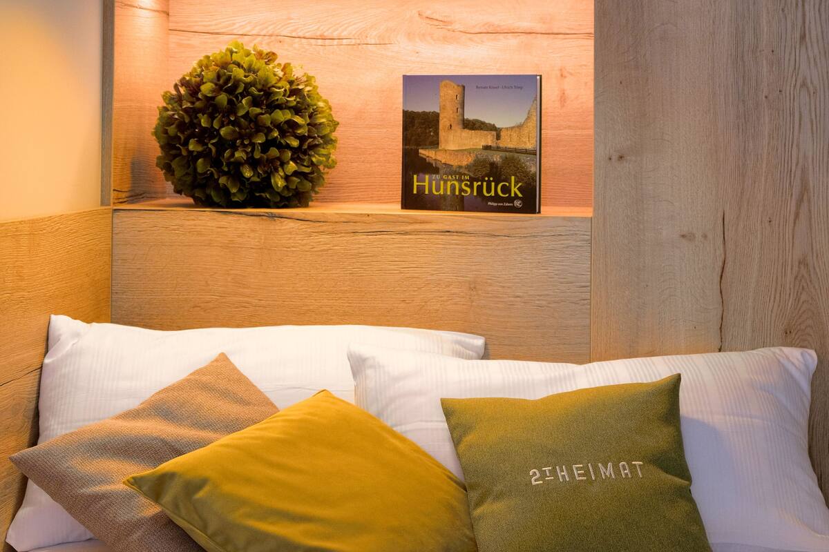 A neatly arranged bed with pillows, a wall-mounted shelf with a book and a decorative plant, and a wall-mounted light fixture.