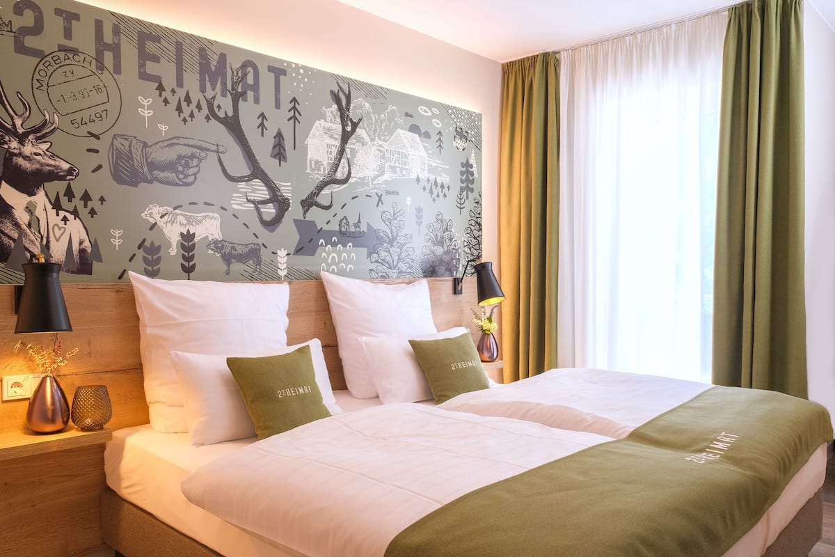A double bed with white and green bedding, a headboard with a wall mural, a nightstand with a lamp, and a window with curtains.