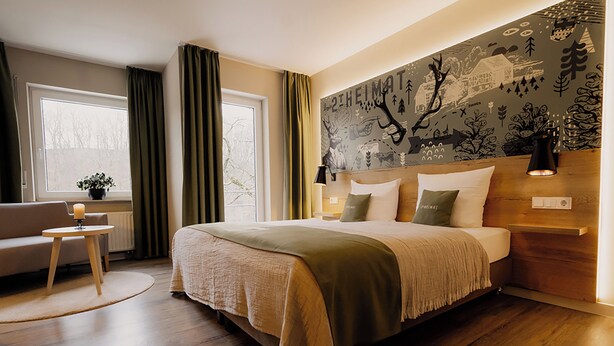 A bedroom with a large bed, a bedside table, a lamp, a window with curtains, and a wall mural.