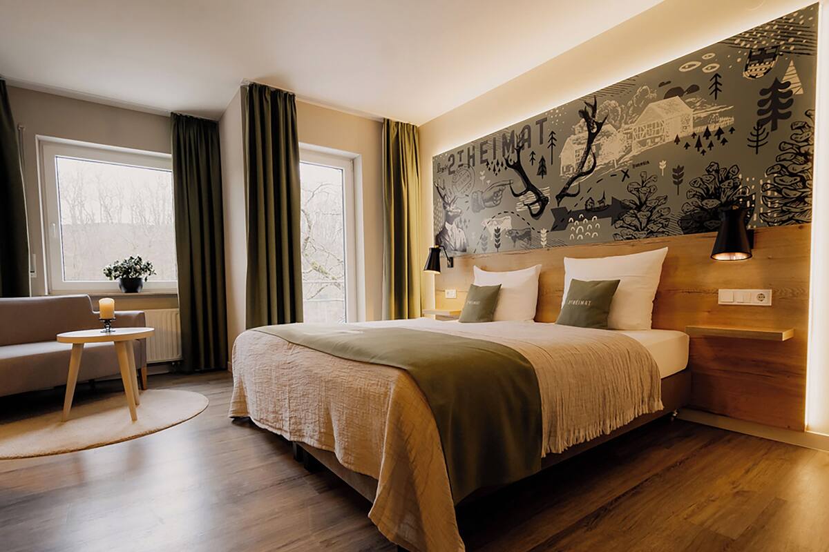 A bedroom with a large bed, a bedside table, a lamp, a window with curtains, and a wall mural.