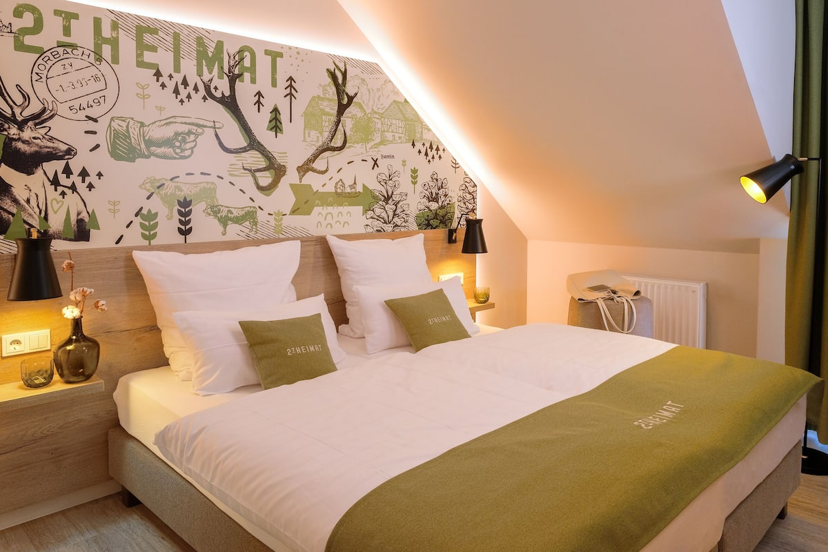 A neatly made bed with white and green bedding, a nightstand with a lamp, and a wall with a nature-themed mural.