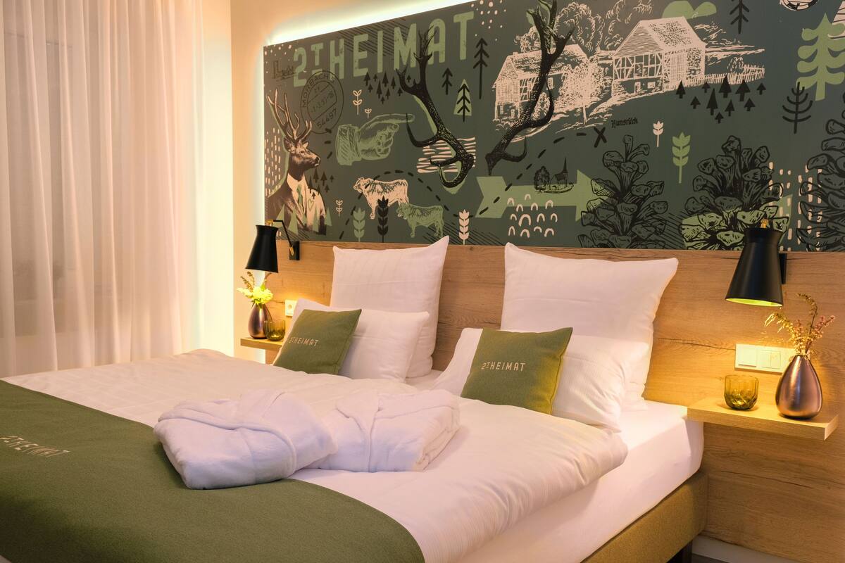 A neatly made bed with white linens and two green pillows, a wooden headboard, and a wall mural with nature and animal illustrations.