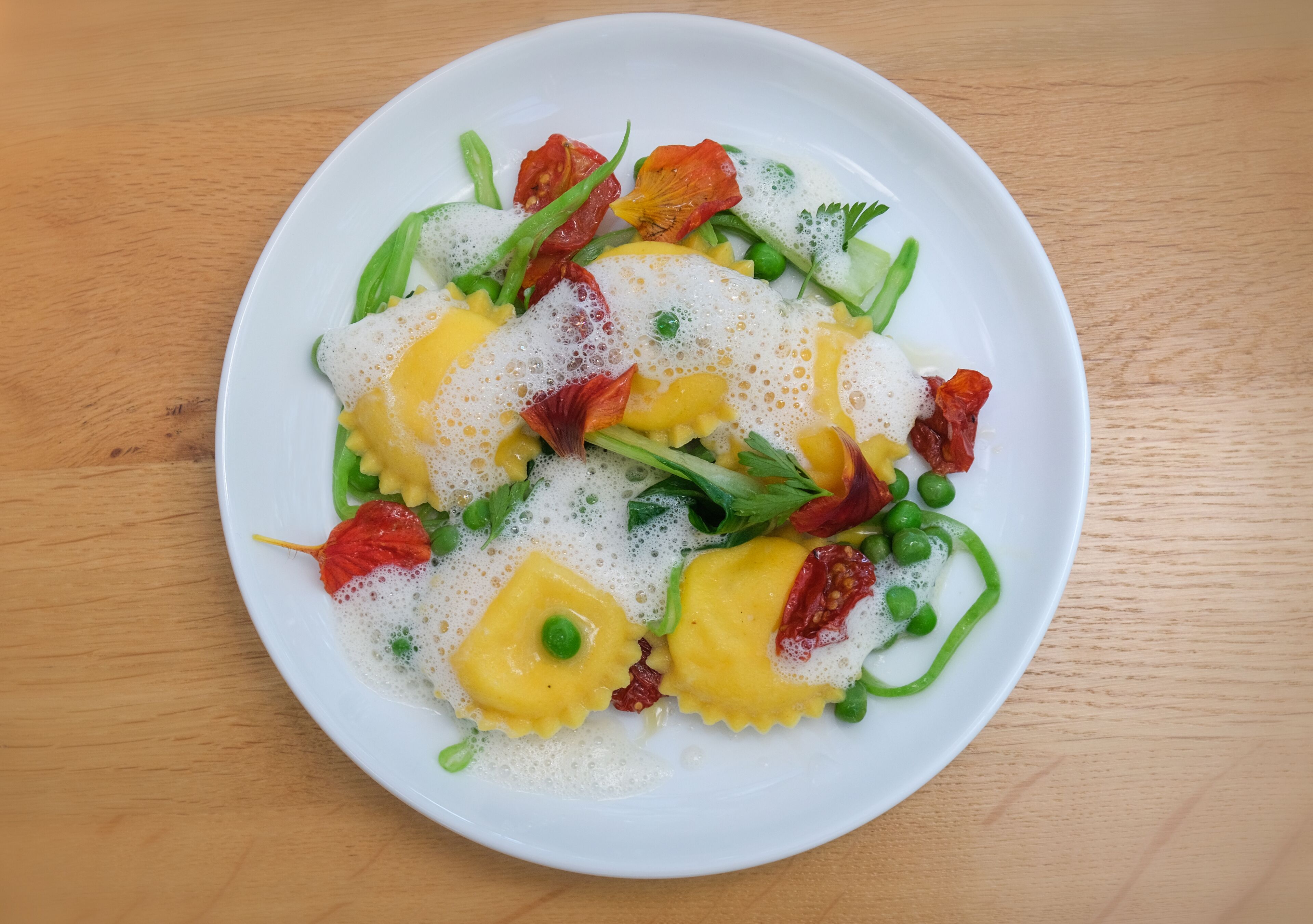 Un plato de ravioles con salsa cremosa, adornado con guisantes verdes, pimientos rojos y hierbas.