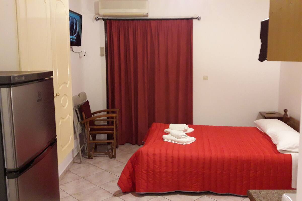 A compact hotel room with a bed, a chair, a small table, and a refrigerator.