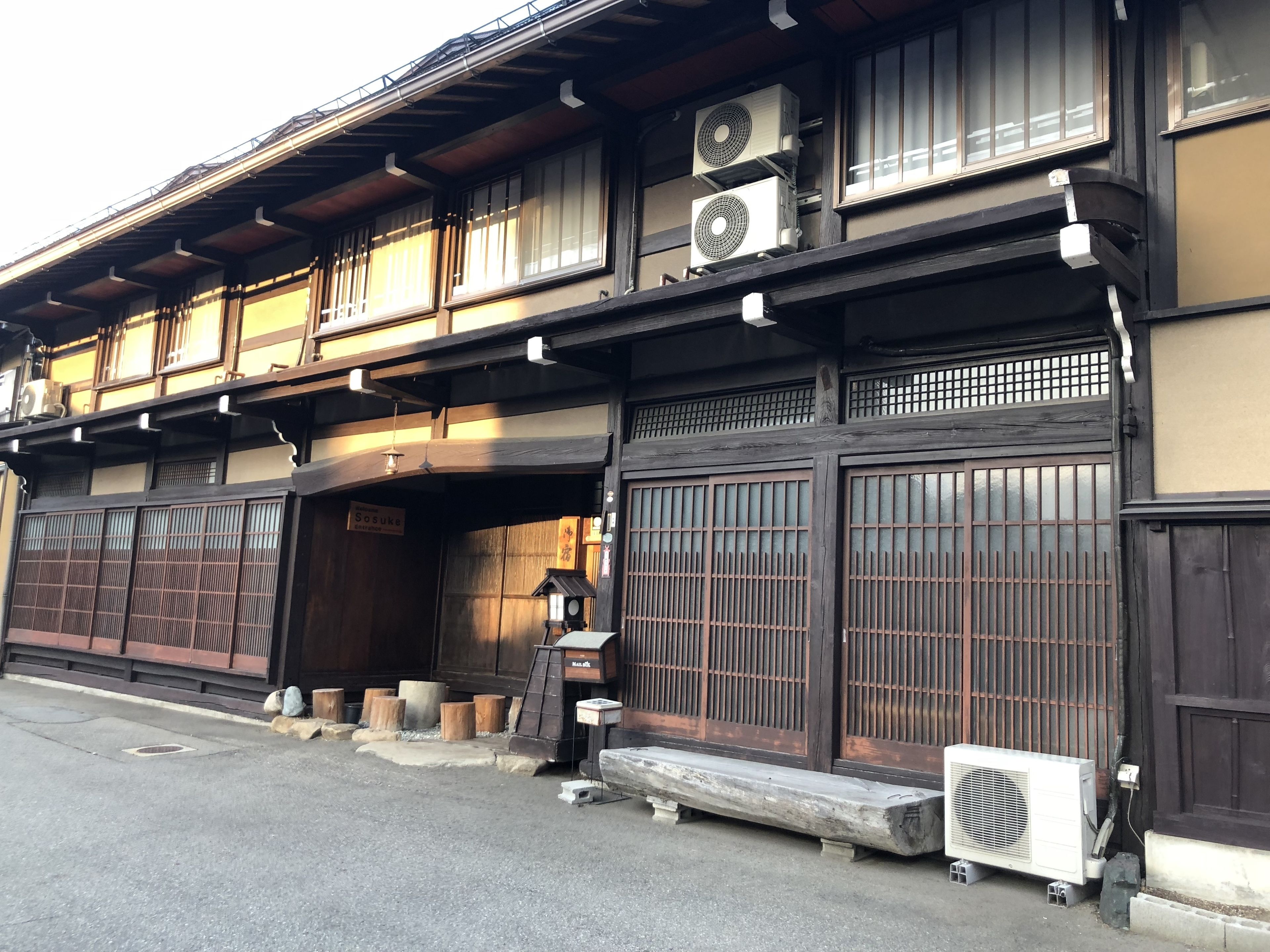 A traditional Japanese building with large wooden sliding doors, a small outdoor seating area, and two air conditioning units on the roof.