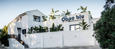 A modern hotel with a white facade, named "Capo blu Hotel & pool bar," featuring a prominent sign and a staircase leading to the entrance.