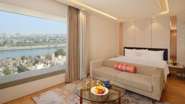 Une chambre d’hôtel moderne avec une grande fenêtre, une table ronde en verre sur laquelle sont disposés des fruits, un canapé et un lit avec une tête de lit.