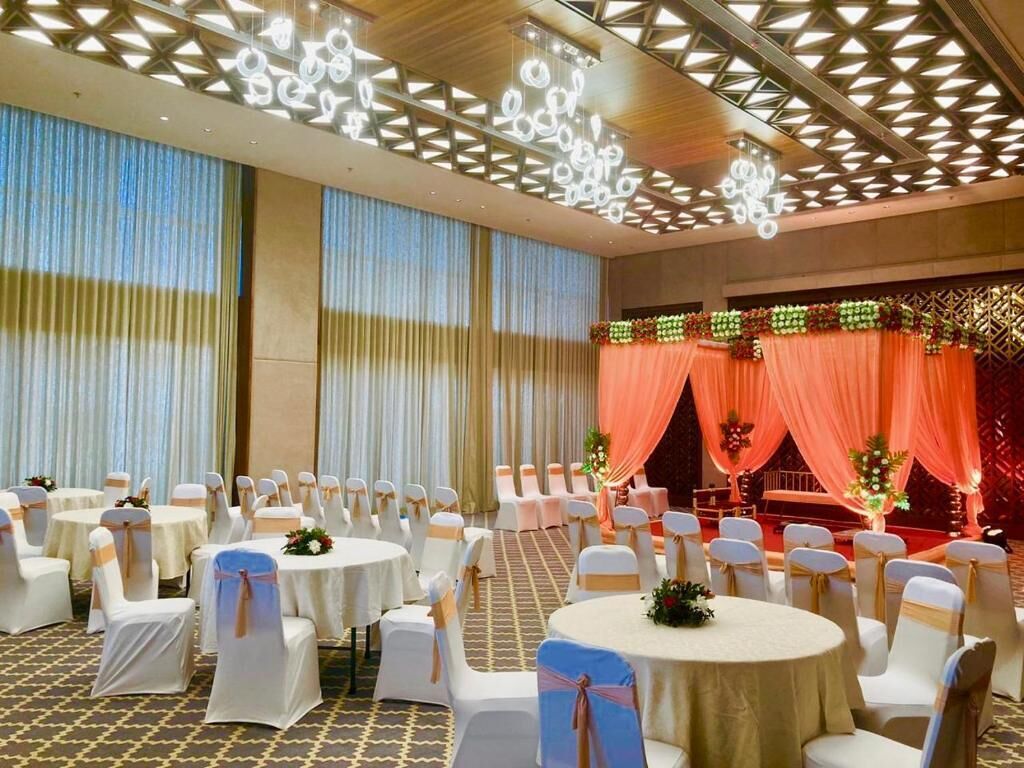A banquet hall with round tables and white chairs, a stage with drapes, and a patterned ceiling.