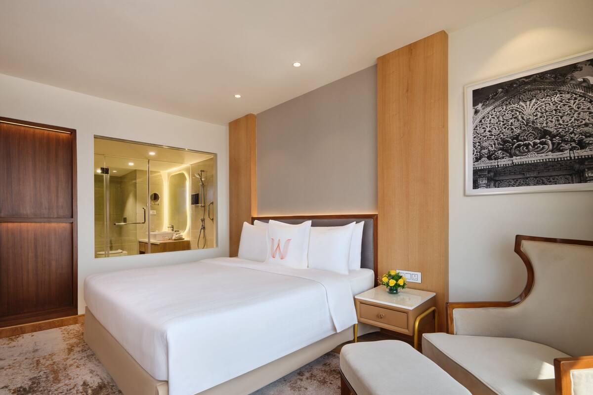 Una habitación de hotel moderna con una cama grande, un armario de madera, un baño con cabina de ducha de cristal y una mesita de noche con un jarrón de flores.