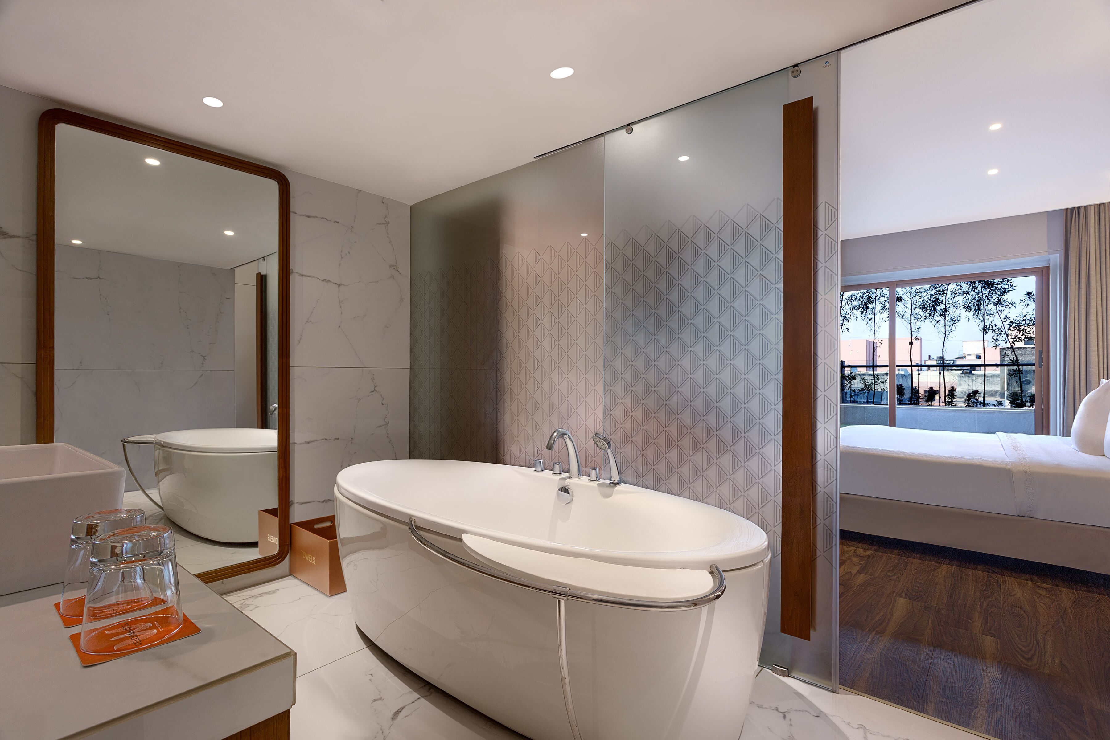A modern bathroom with a freestanding bathtub, a large mirror, and a view of the outside through a glass door.