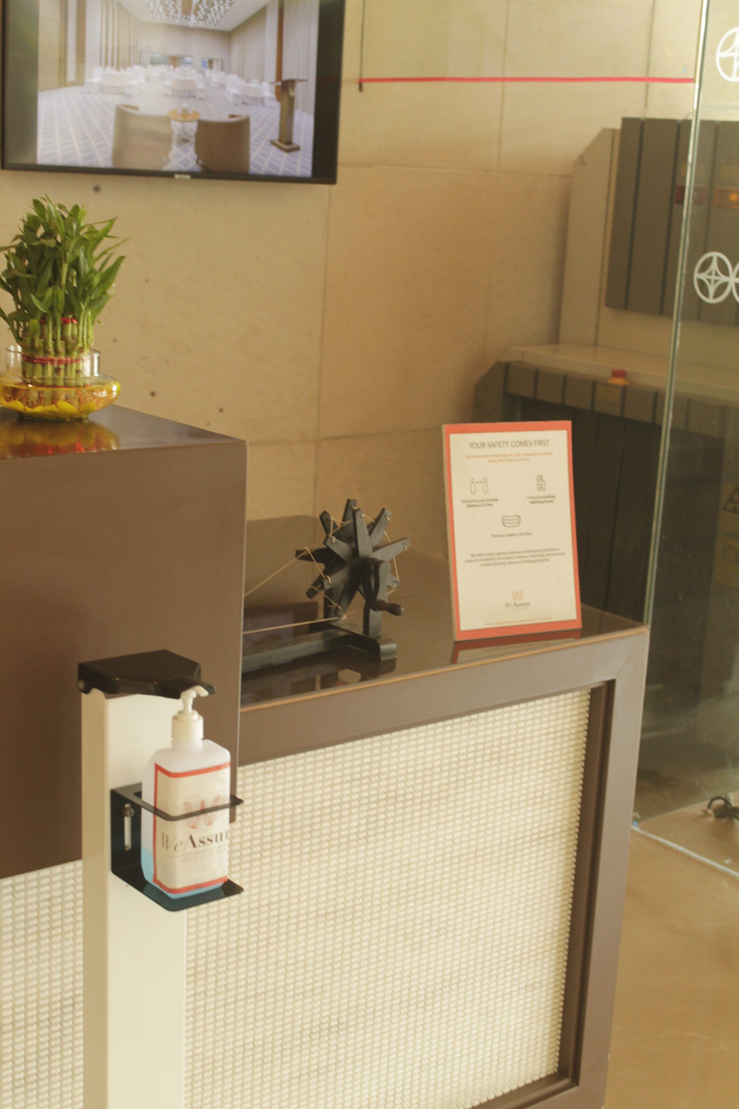 A reception desk with a television displaying a room layout, a potted plant, a hand sanitizer dispenser, and a safety notice.