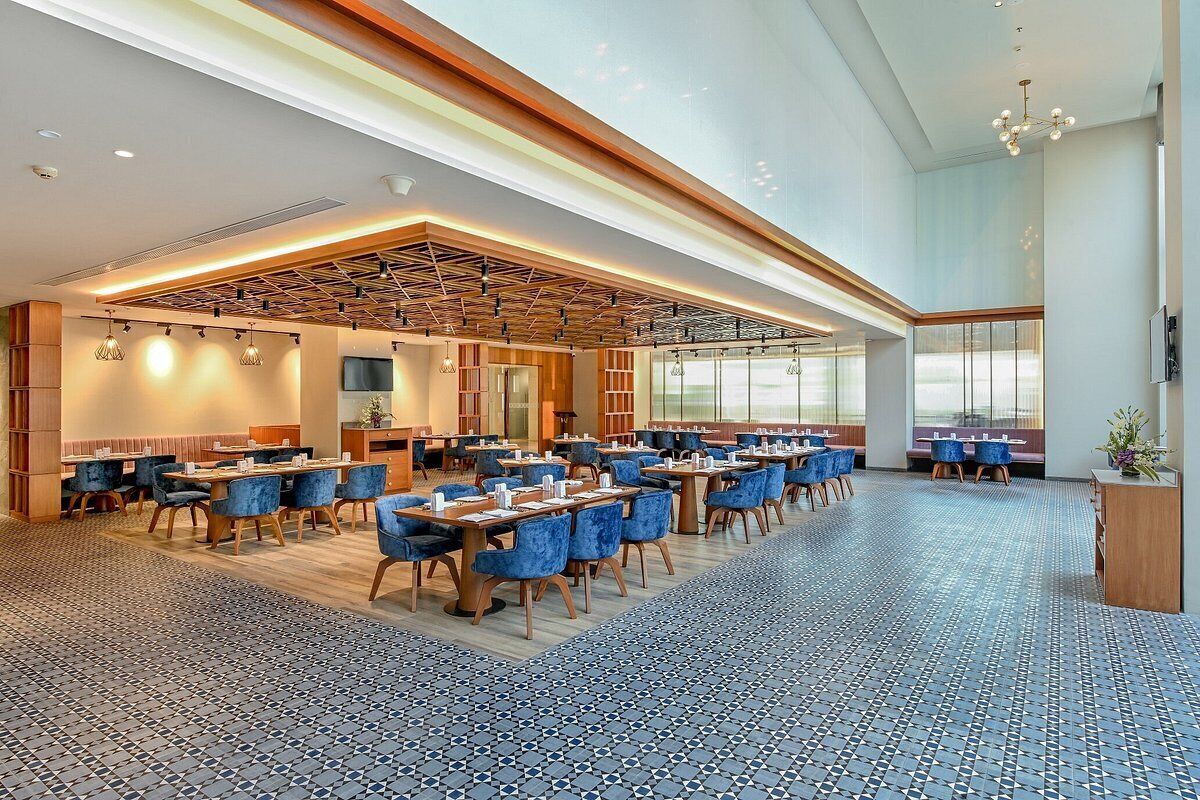 A spacious hotel dining area with multiple tables and chairs, a modern ceiling design, and large windows providing natural light.