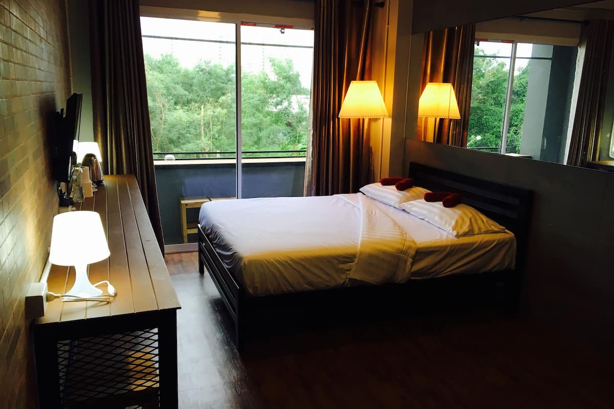 A hotel room with a bed, bedside lamps, a wooden desk, and a view of greenery through large windows.