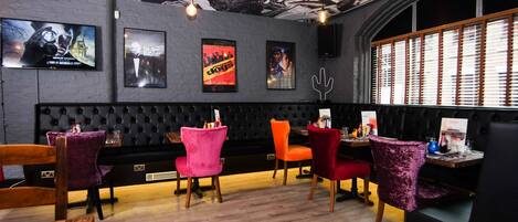 Un interior de restaurante con un asiento acolchado tipo cabina negro, sillas coloridas y carteles de películas enmarcados en la pared.