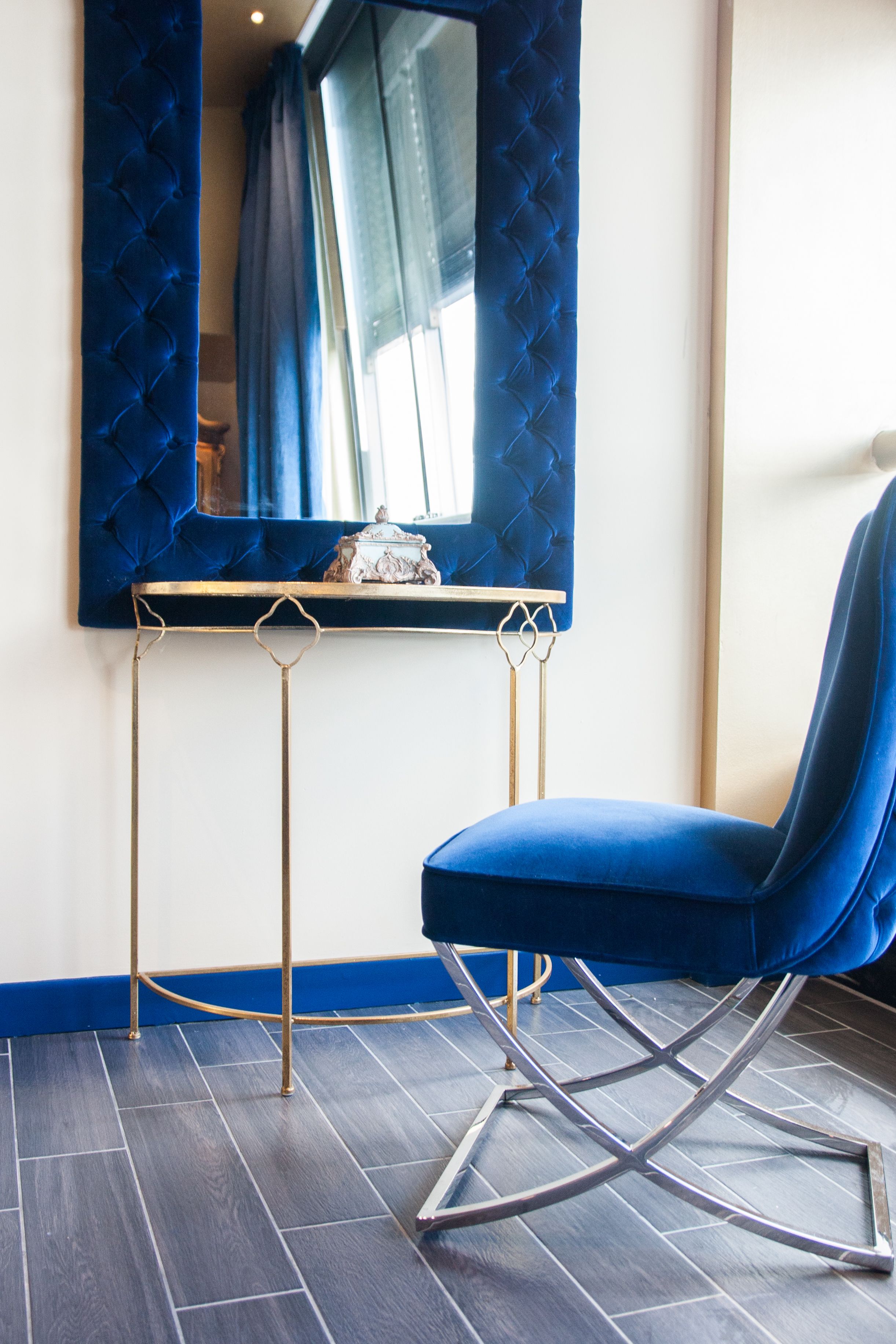 A room with a blue upholstered chair, a small table with a decorative item, and a mirror with a blue curtain.