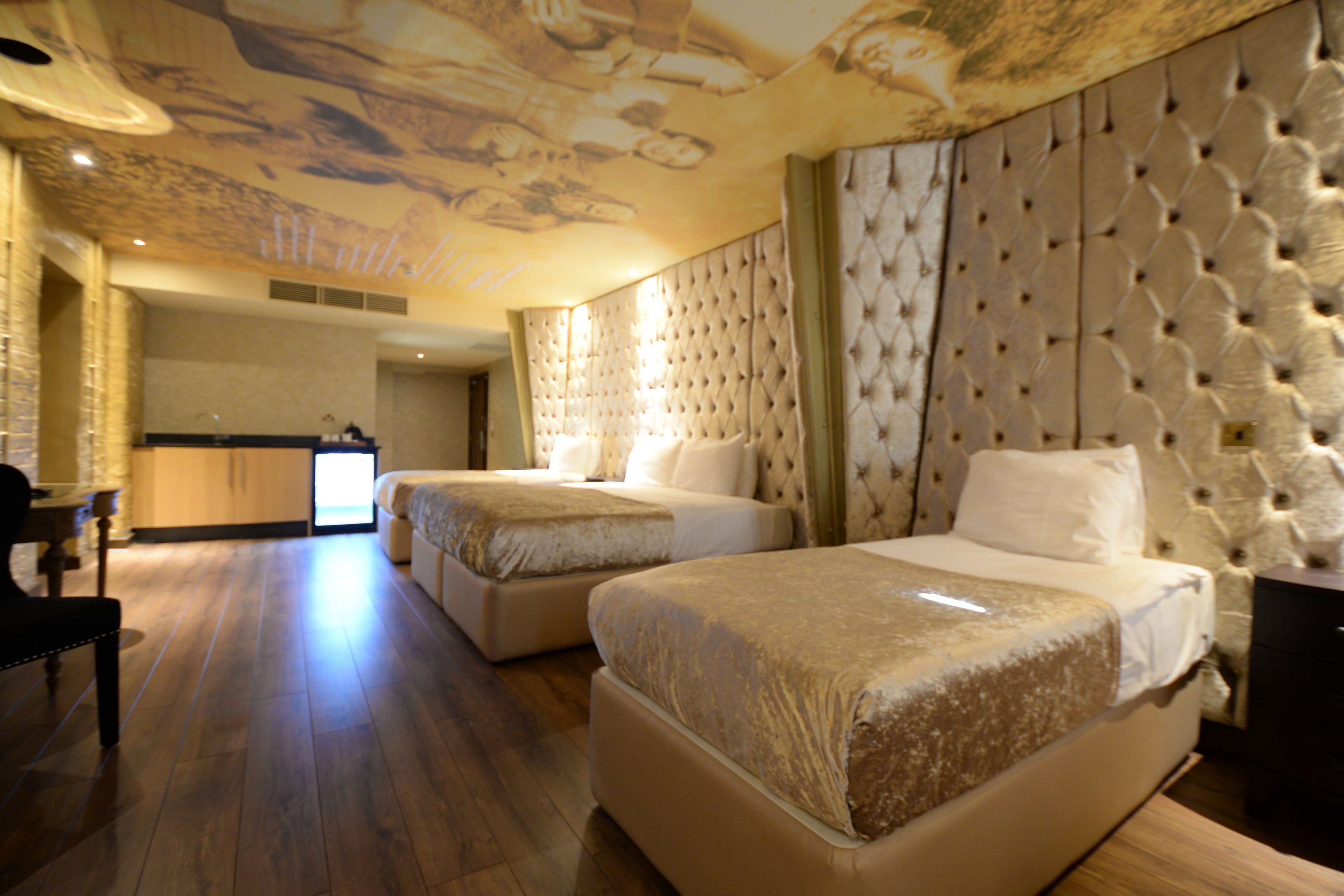 A hotel room with two beds, a wooden floor, a large mural on the ceiling, and a view of the kitchen.
