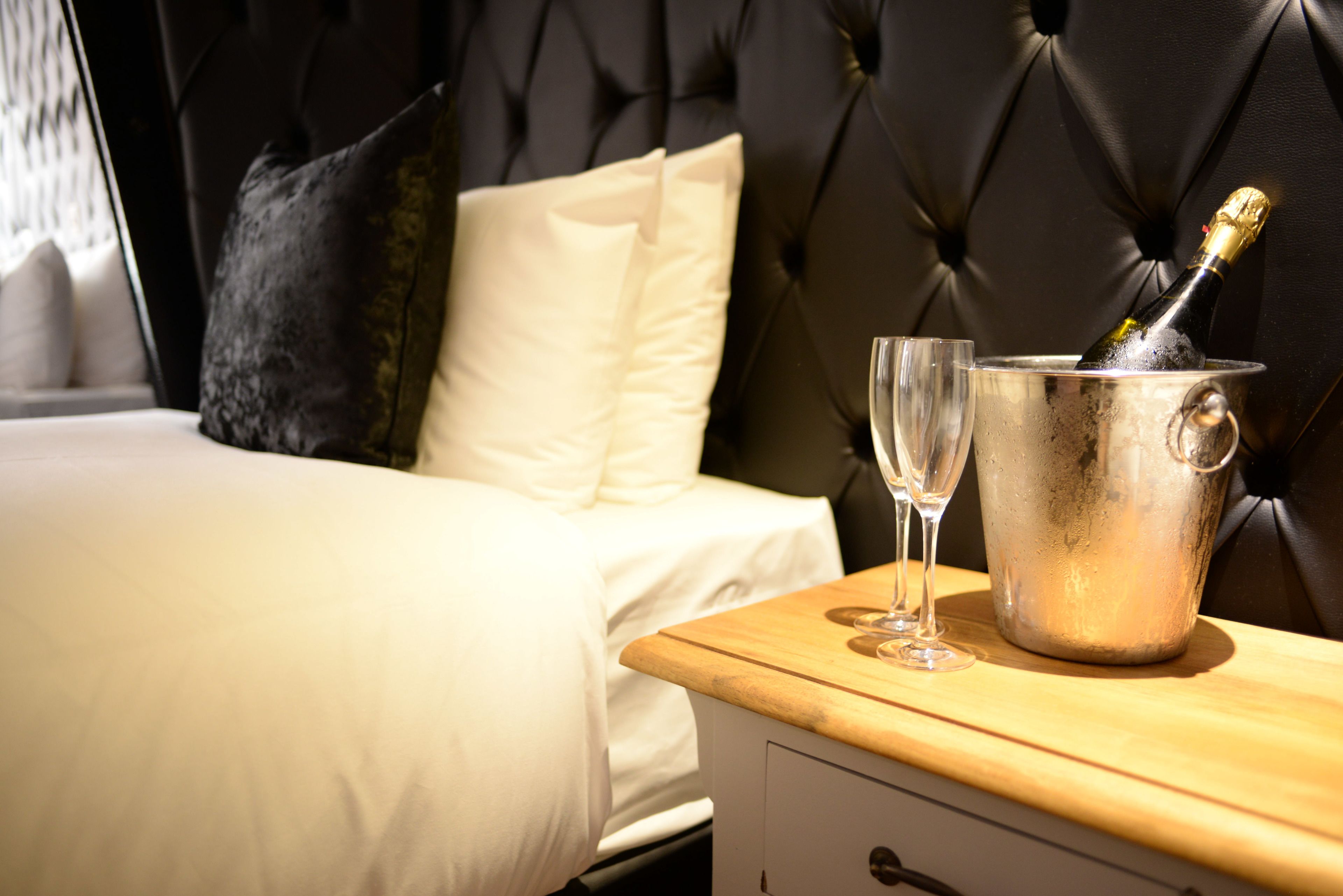 A bed with white linens, a black tufted headboard, and a wooden bedside table with a champagne bottle and two glasses.