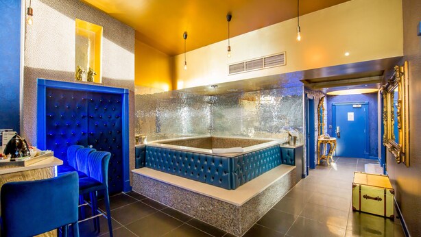 Une salle de bain luxueuse avec une grande baignoire, un coin salon doté de fauteuils recouverts d’un tissu bleu, et un miroir orné d’un cadre travaillé.