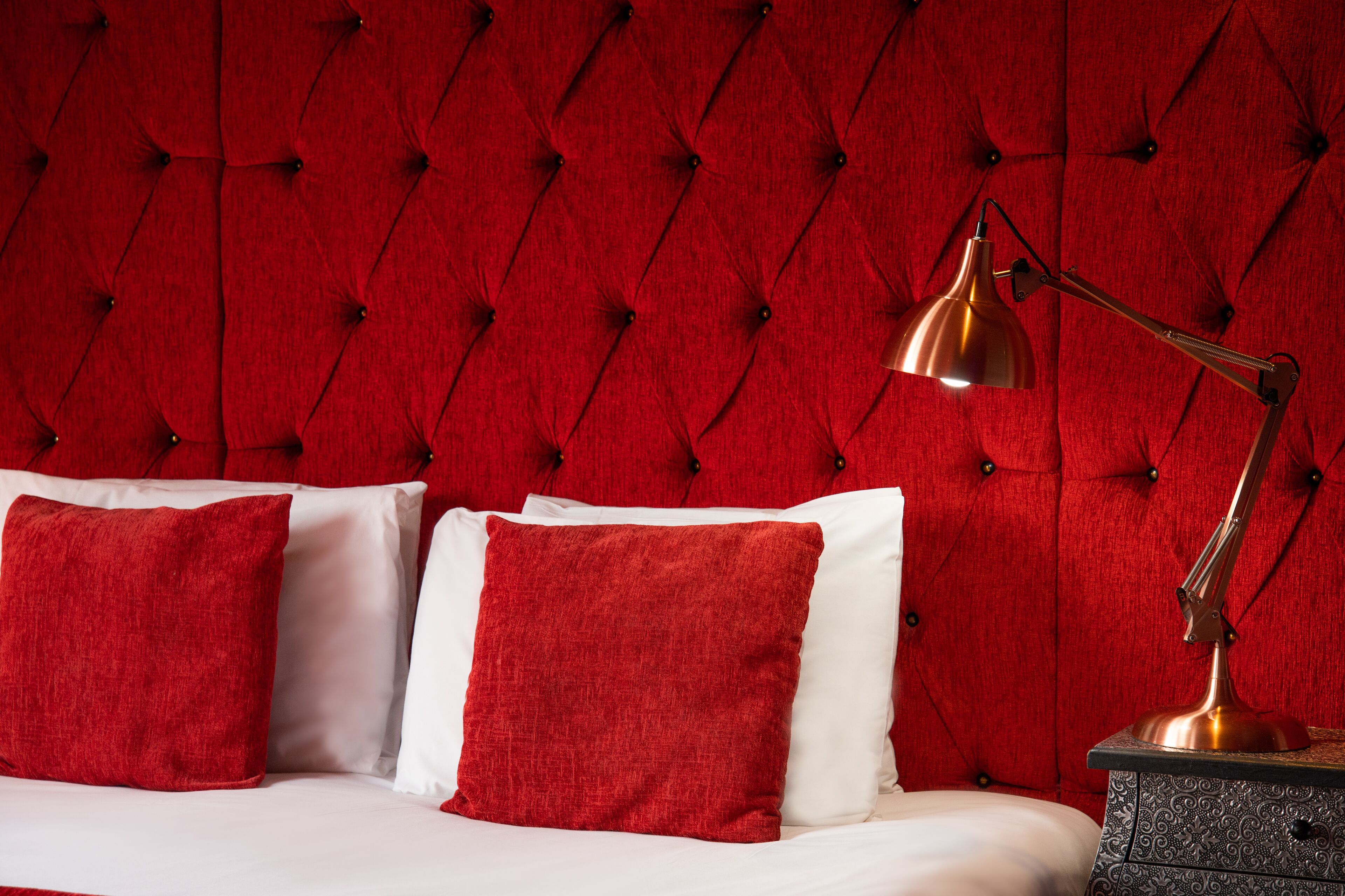 Un lit avec des oreillers rouges et blancs, une tête de lit rouge à motifs et une lampe de bureau en laiton.