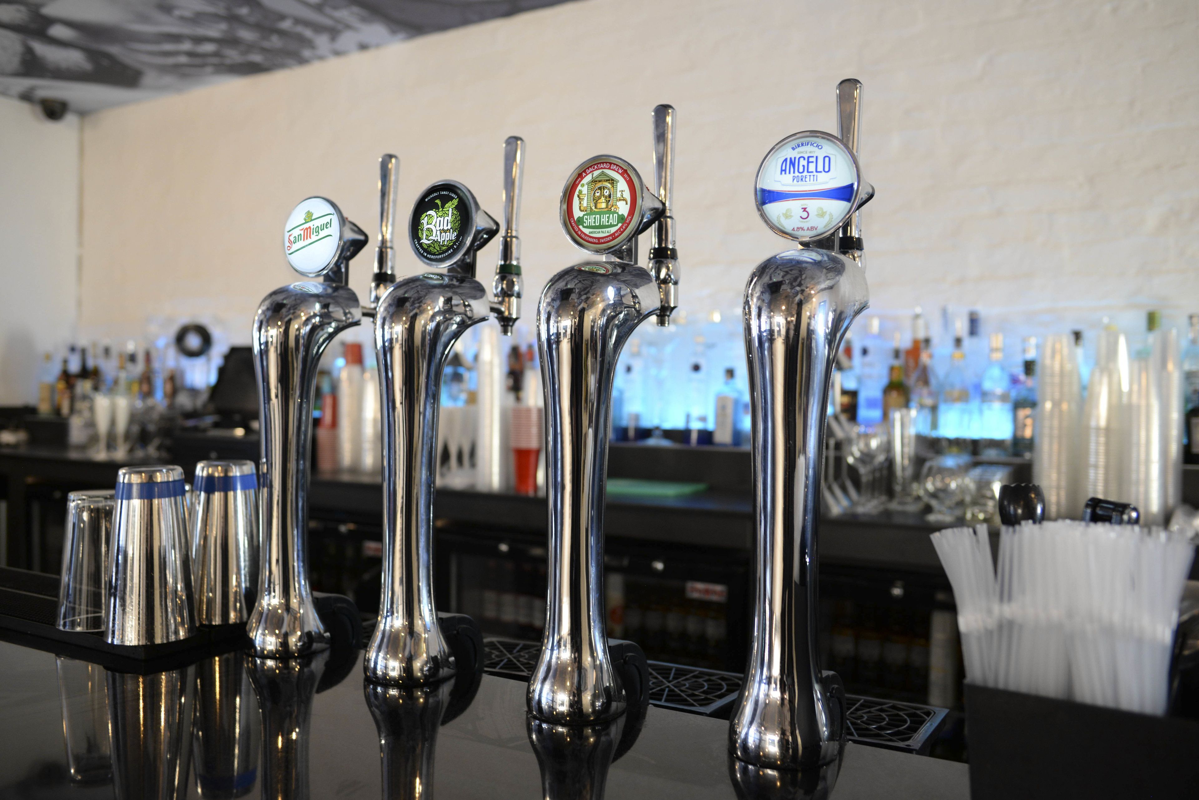 A row of beer taps with different brand labels, a bar counter, and various bottles and glasses in the background.