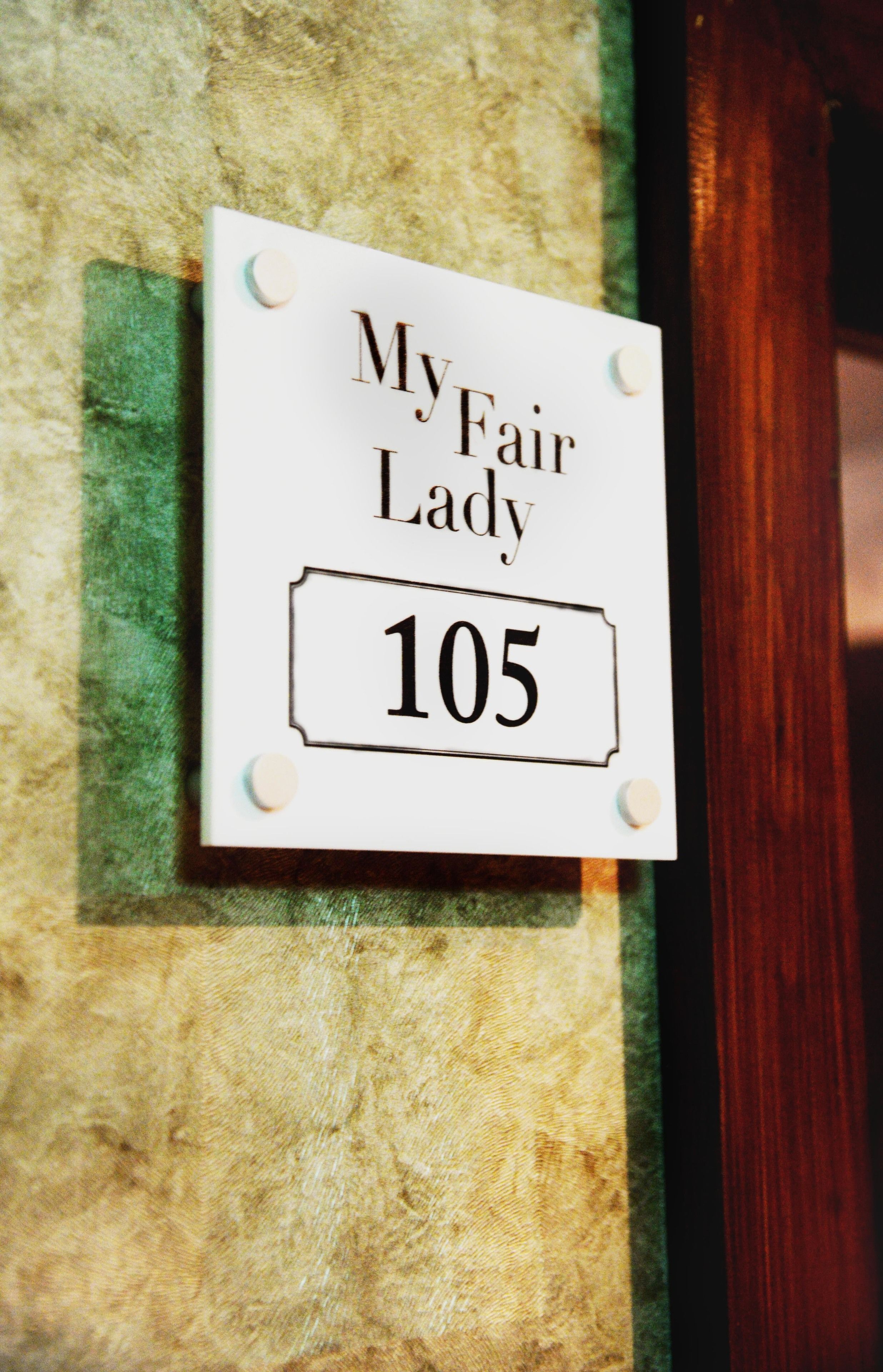 Tekstillä ”MyFairLady 105” varustettu kyltti on kiinnitetty kuvioituun seinään.