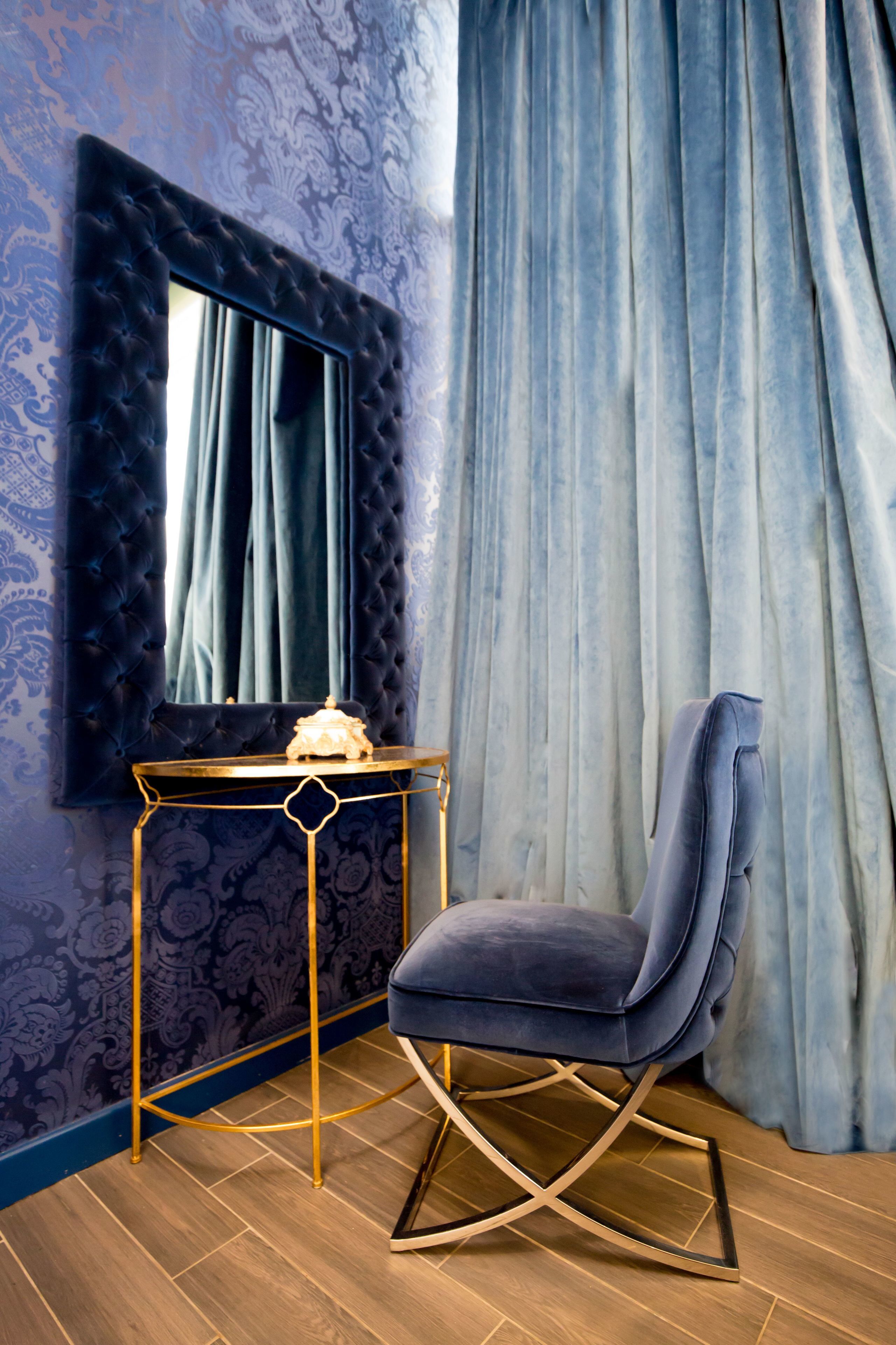 Una habitación con un cabecero capitonado, una mesita consola redonda dorada, un sillón de terciopelo azul y un papel tapiz estampado.