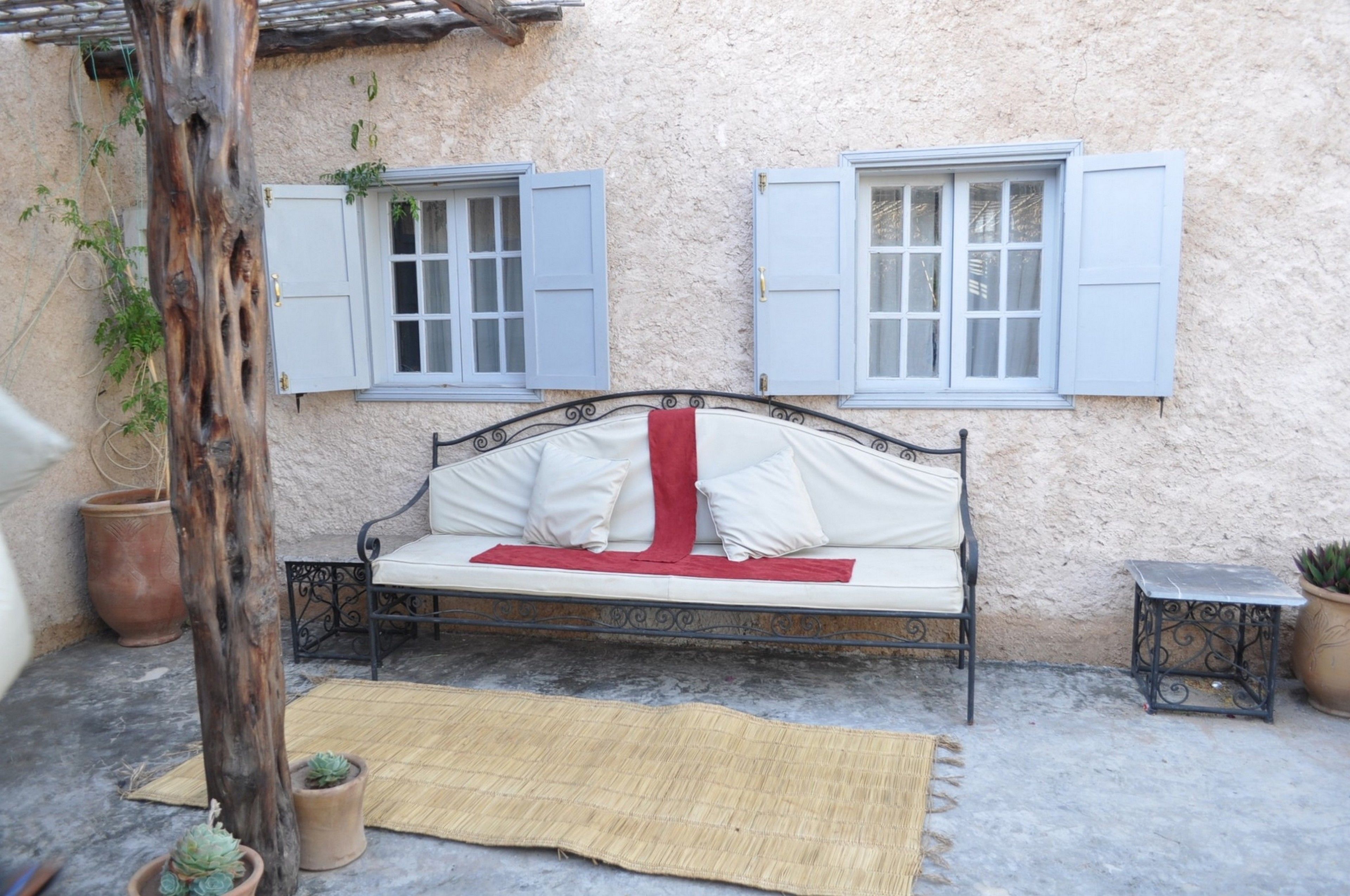 A bed with white bedding and a red throw, a small table, and two windows with blue shutters.