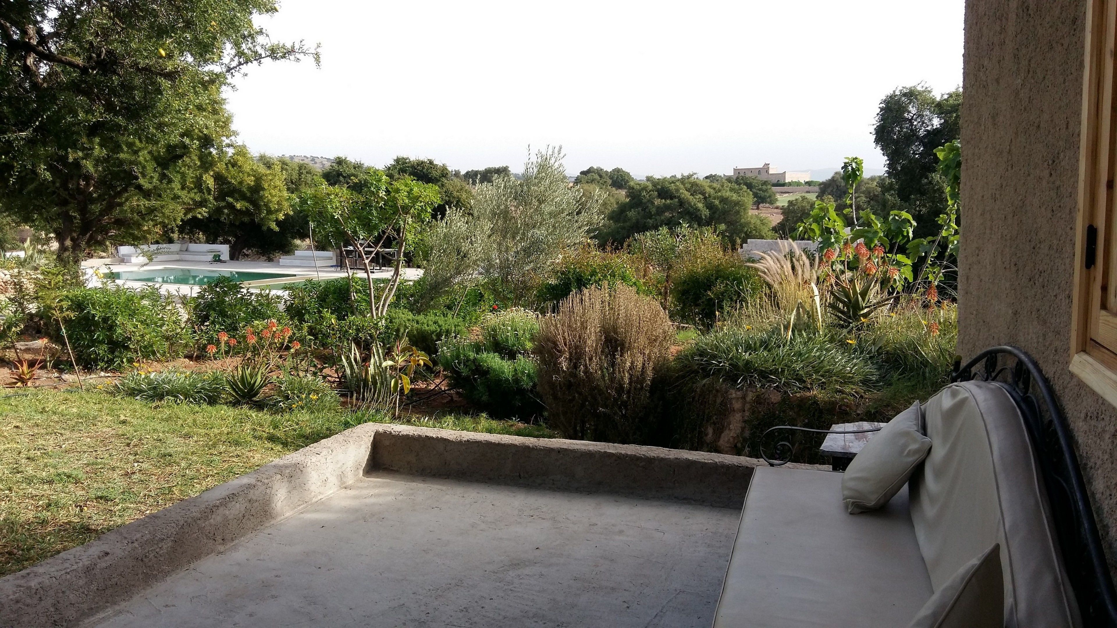 A patio with a concrete floor, a white cushioned bench, and a view of a swimming pool and greenery.