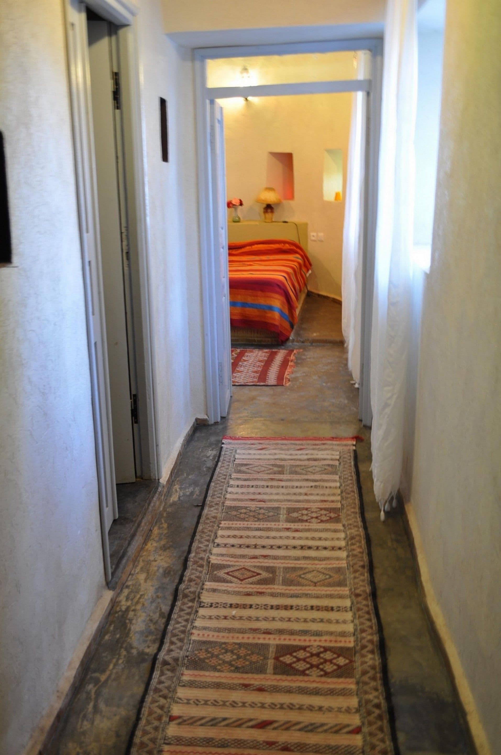 Ein schmaler Flur mit gemusterten Teppichen führt zu einem Schlafzimmer mit Bett und Nachttisch.