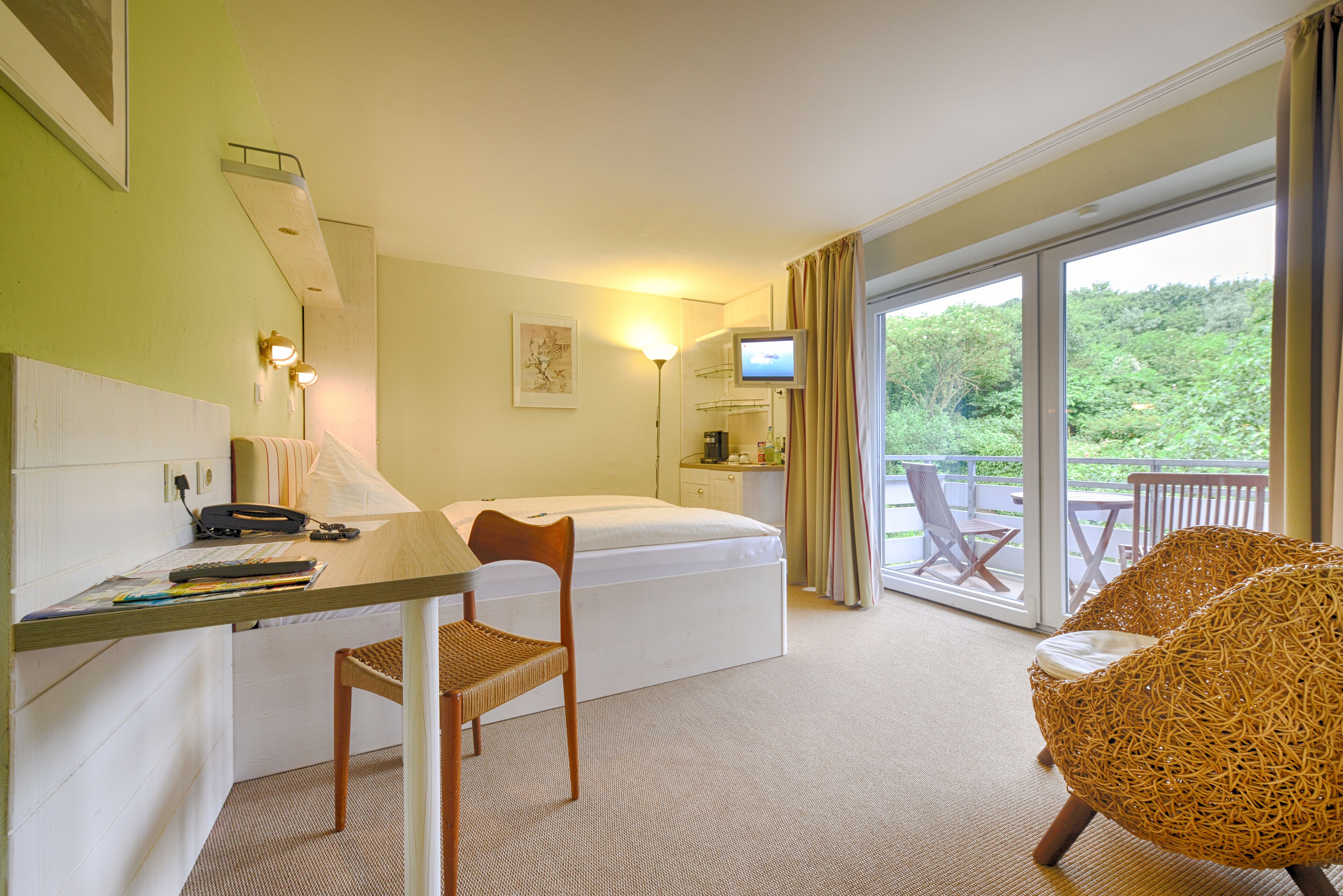 A hotel room with a bed, a desk with a chair, a television, and a balcony with a wicker chair.