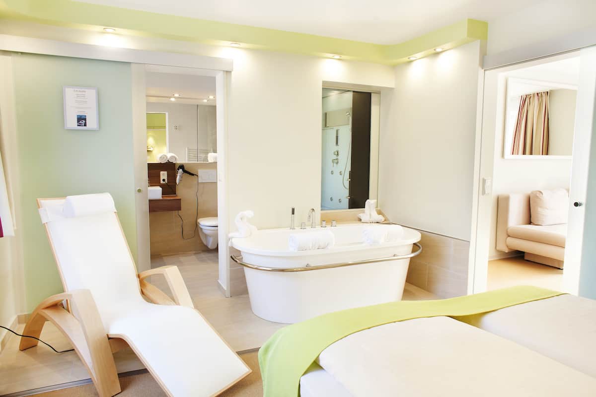 Une salle de bain moderne avec une baignoire, une douche et des toilettes. On y trouve une chaise et un lit recouvert d’une couverture verte.