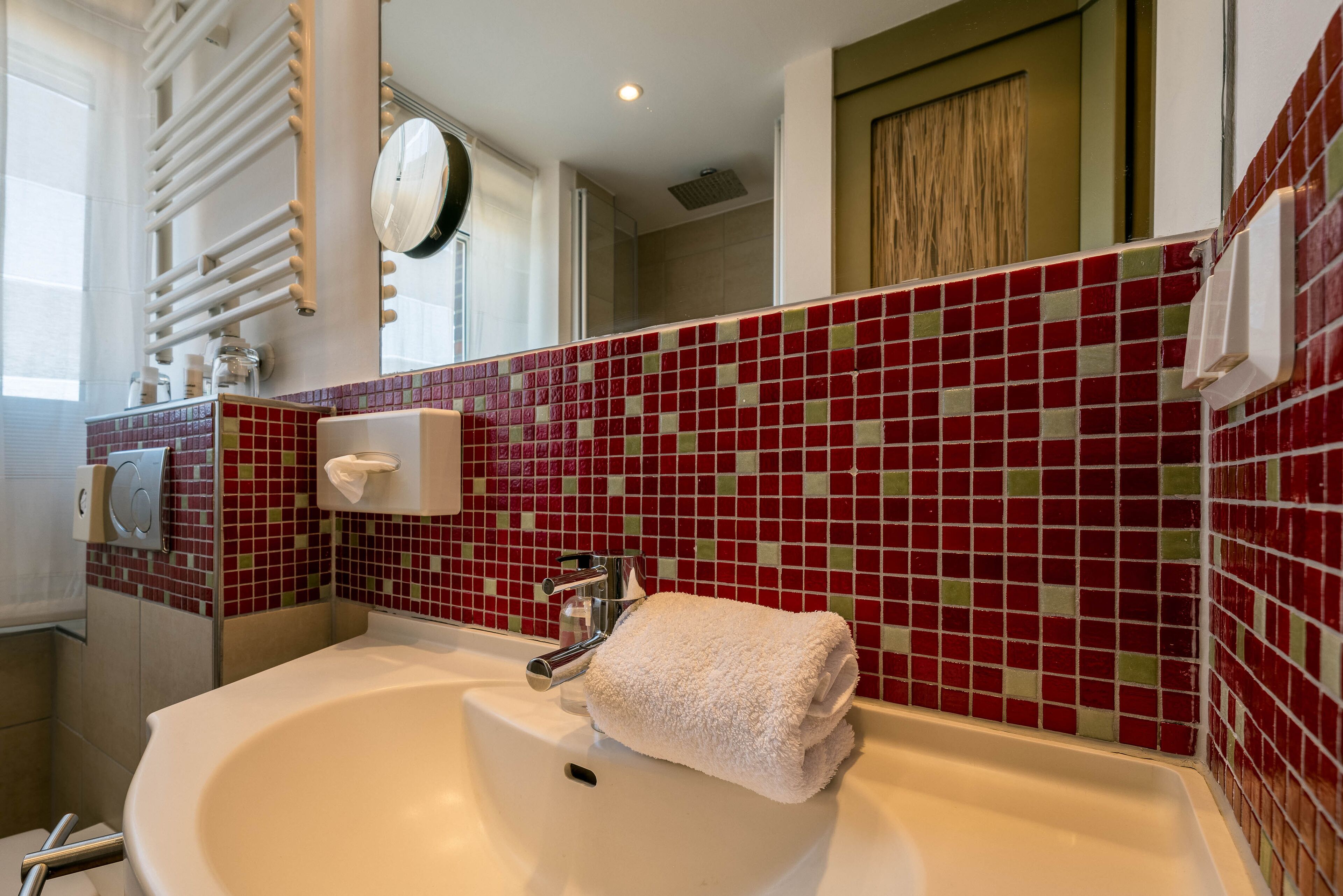Un baño con un lavamanos blanco, mosaicos de colores rojo y verde, una toalla y un espejo.