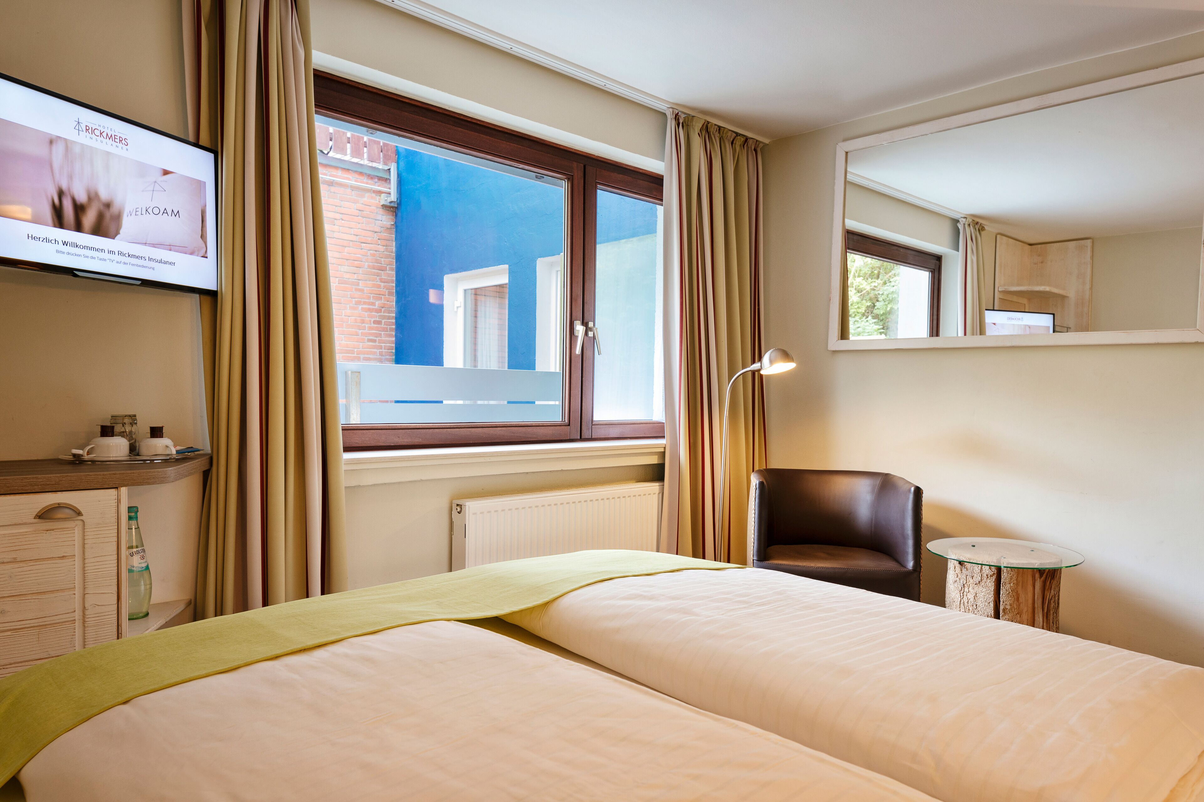 A hotel room with two beds, a TV on the wall, a window with curtains, a chair, and a small table.