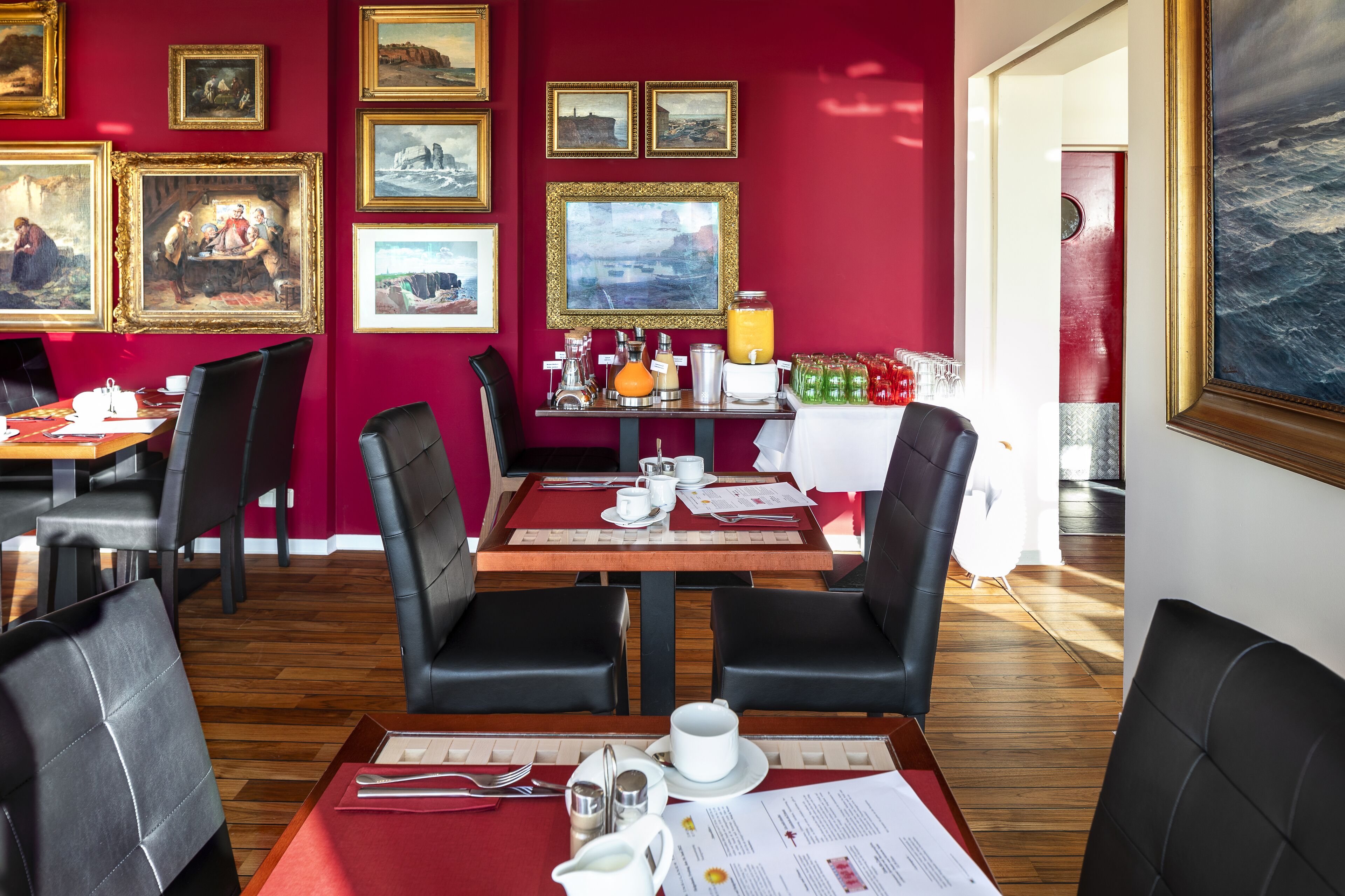 A dining area with red walls, multiple framed artworks, and wooden tables with black leather chairs.