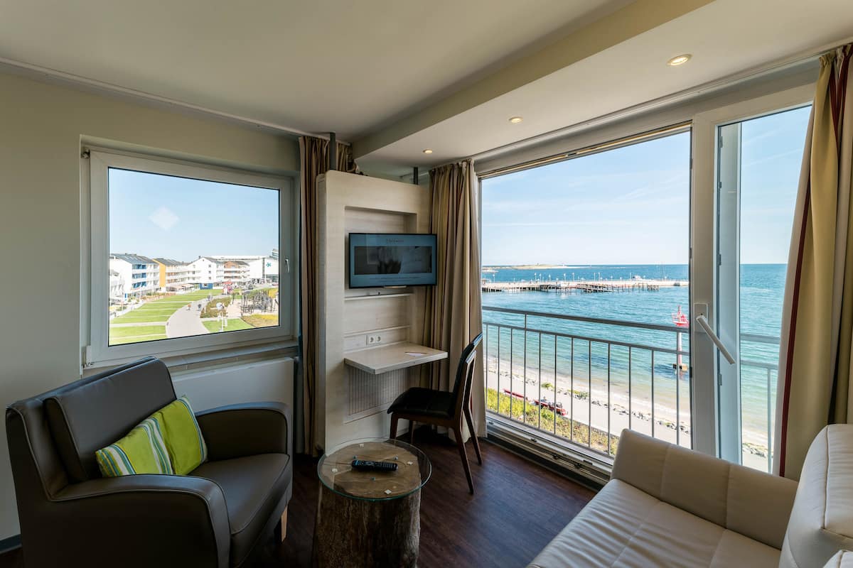 Une chambre d’hôtel avec un balcon, un téléviseur fixé au mur, un canapé, un fauteuil et une petite table.